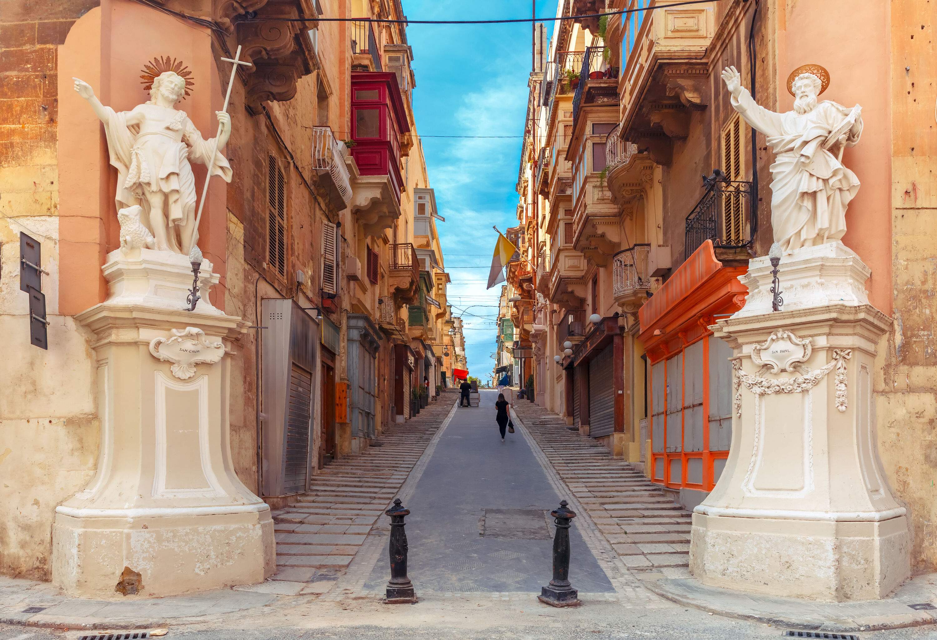 A steep path between ancient buildings guarded by two religious sculptures and bollards on each side.