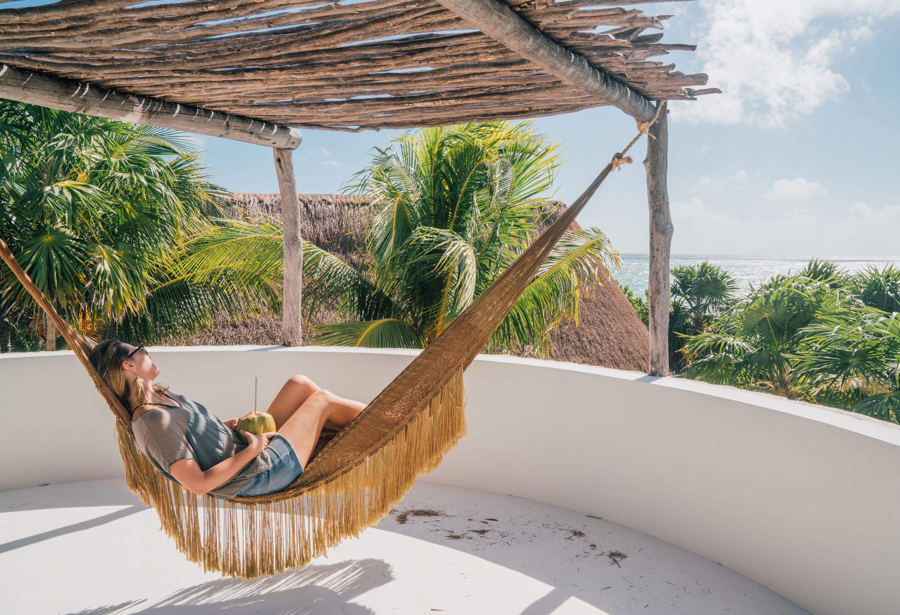 A blonde woman is holding a coconut fruit on a hammock surrounded by palm trees.