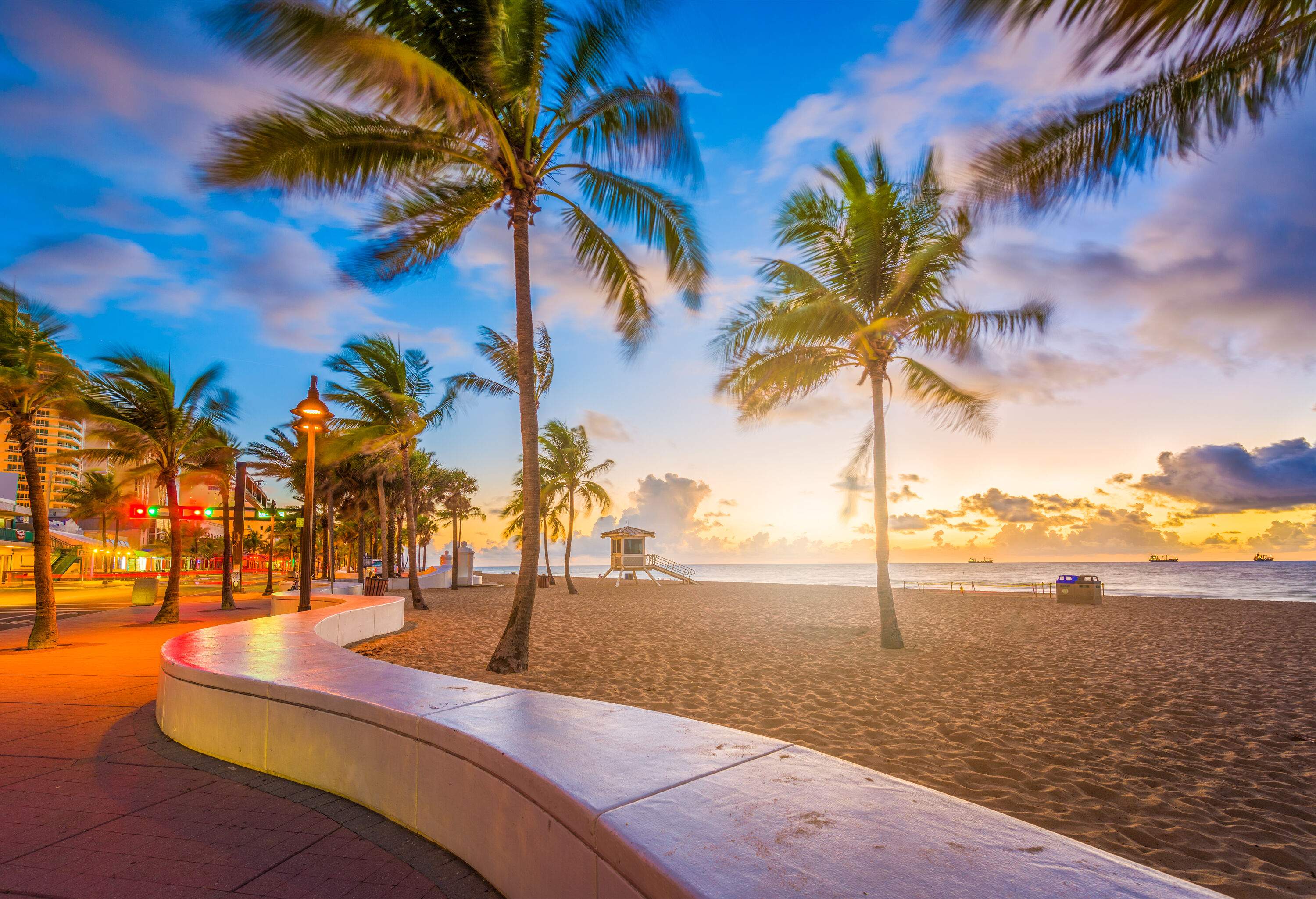 A lifeguard tower and palm trees stand tall on the empty sandy shore alongside a promenade against the scenic sunset.