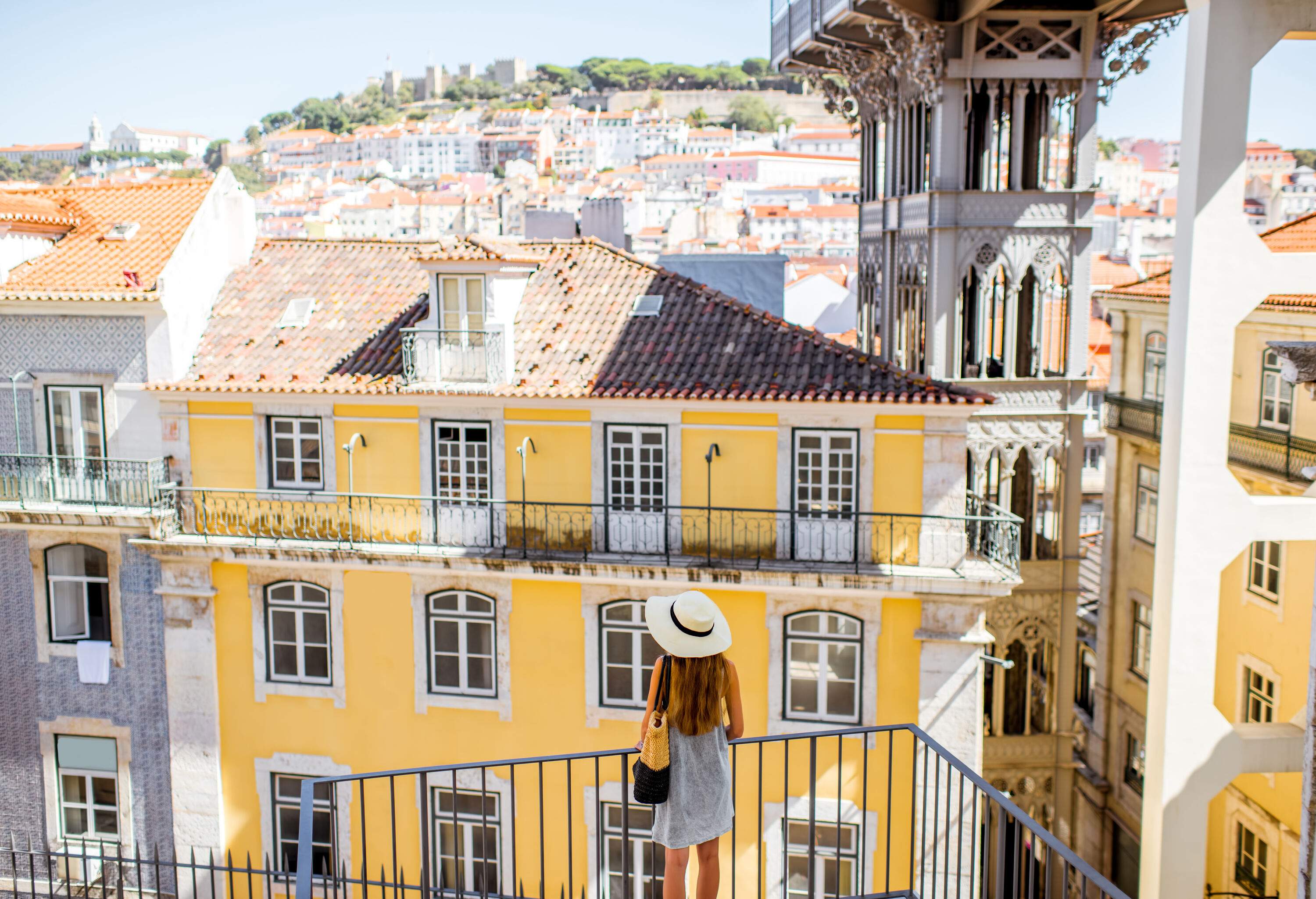 A long-haired blonde woman stands on a balcony with a view of compact buildings ascending the hill.