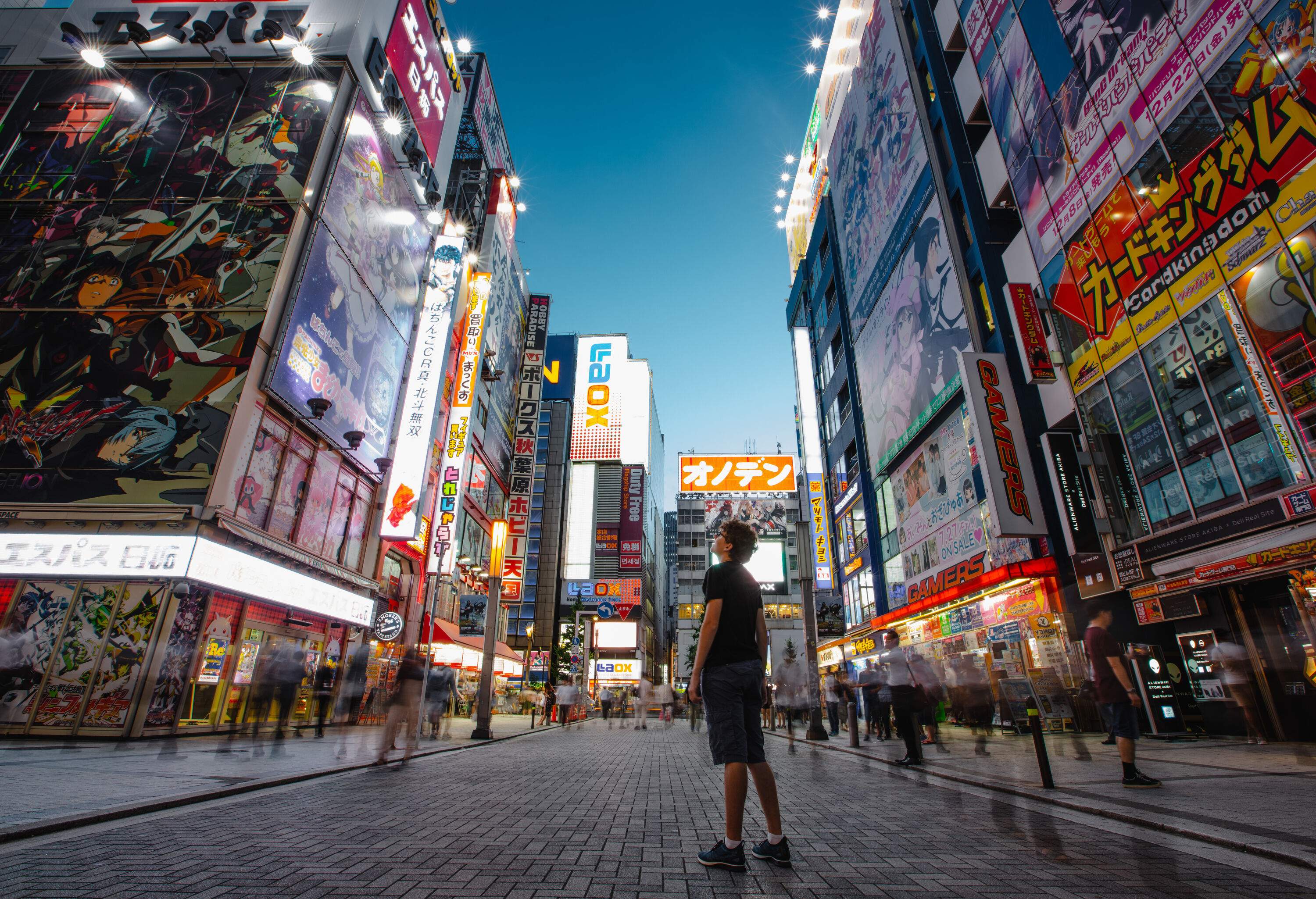 A young man is surrounded by the frenetic energy of a city in motion, with people whizzing by and a riot of colourful advertisements plastered across the building walls.