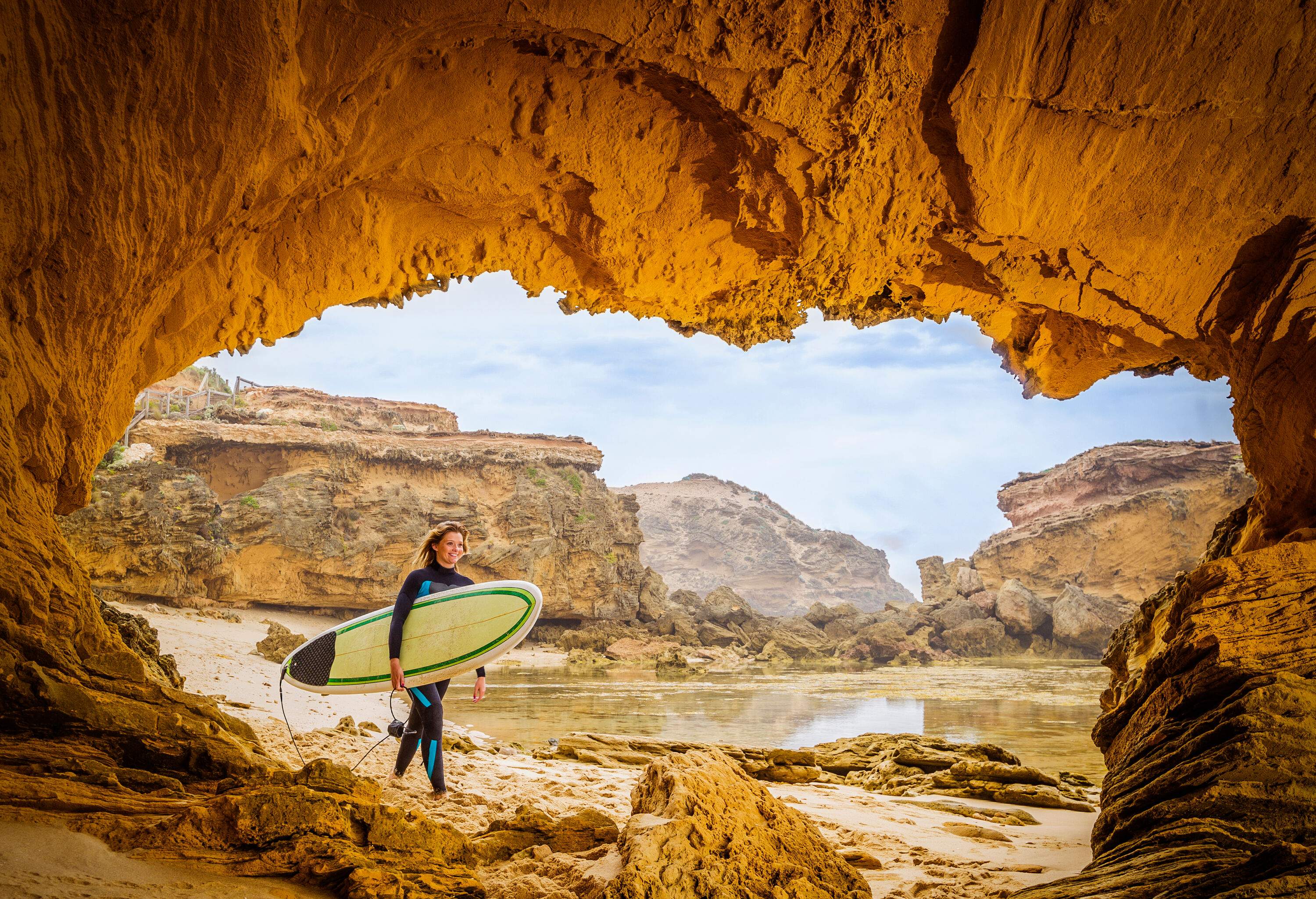 A young female surfer in a wetsuit carrying a surfboard as she walks by a cave's opening.
