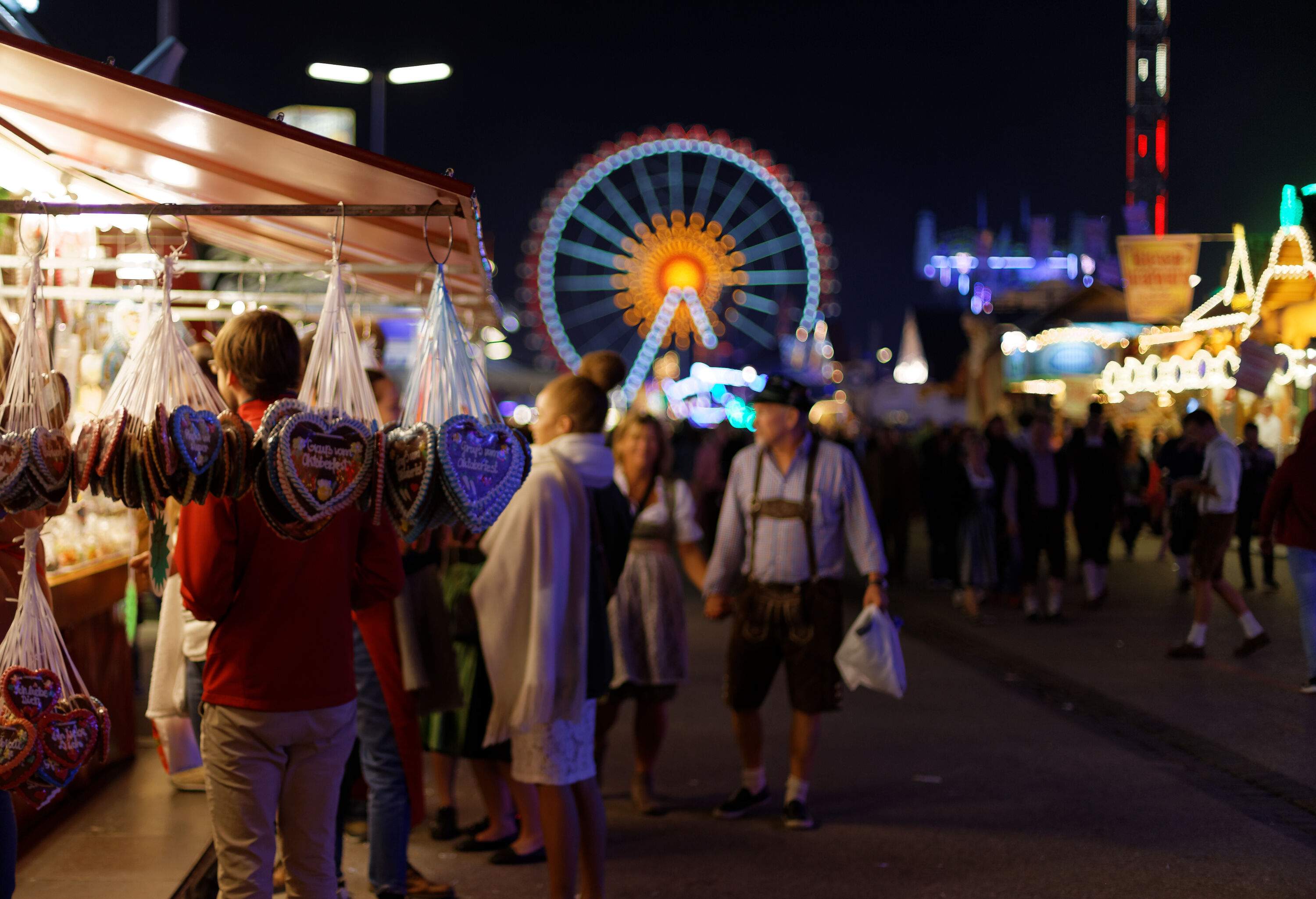 People flocked to a festival in the middle of various booths with a lit Ferris wheel in the background.