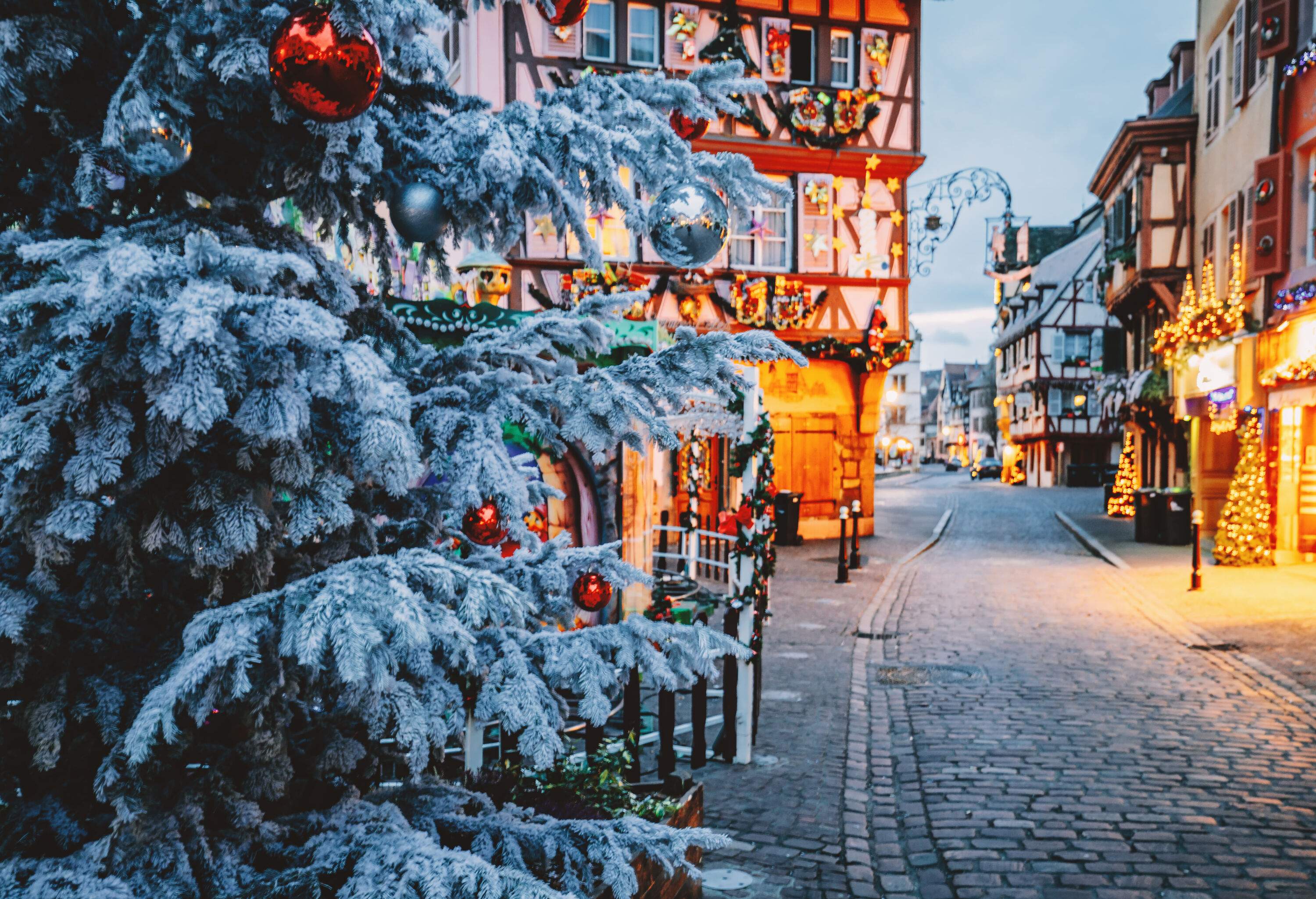A cobbled street lined with half-timbered houses decorated with various colourful Christmas ornamentals and Christmas lights lightly covered in snow.