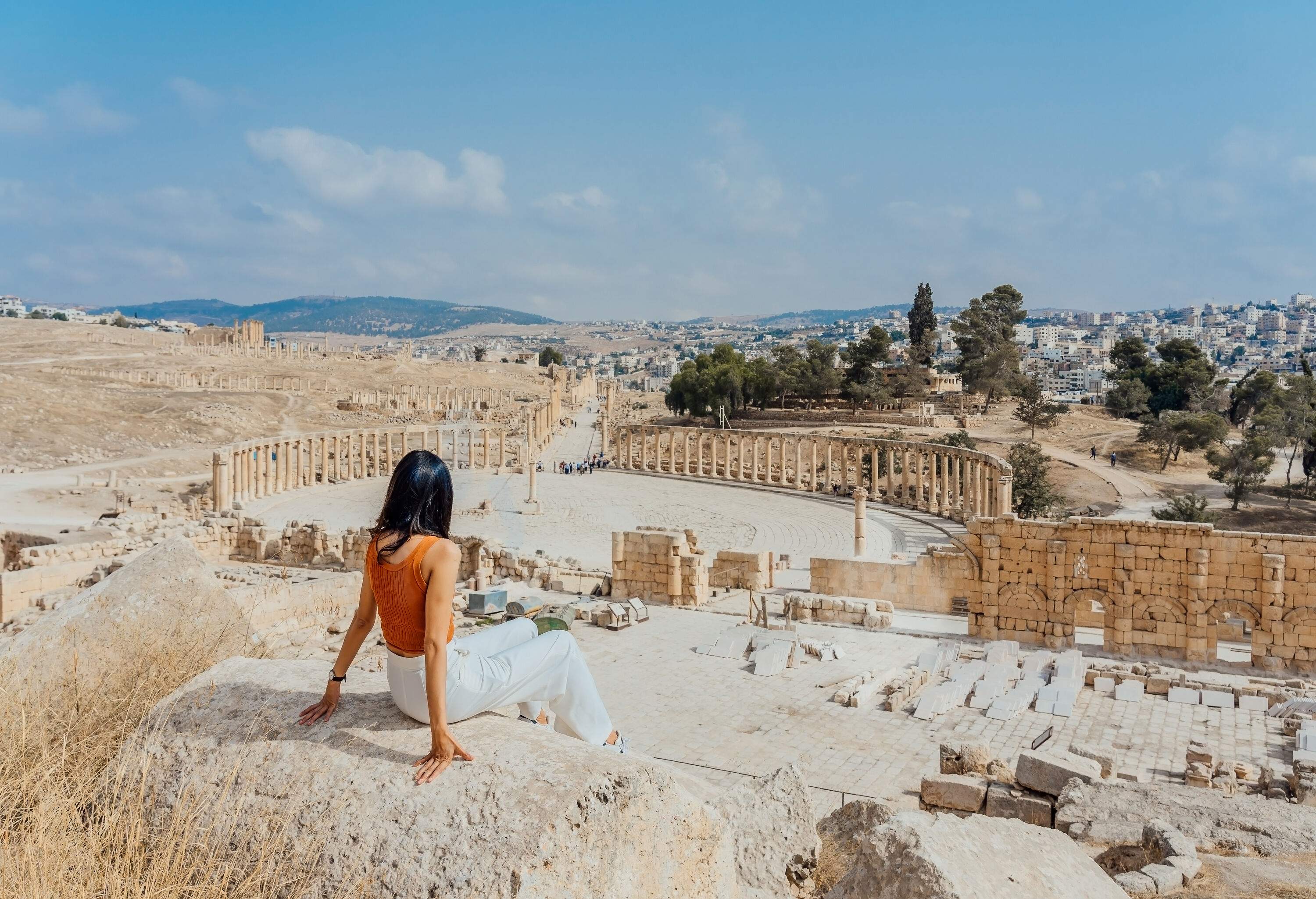 A lady tourist sits on a boulder overlooking the ruins of an impressive and well-preserved plaza circled by columns.