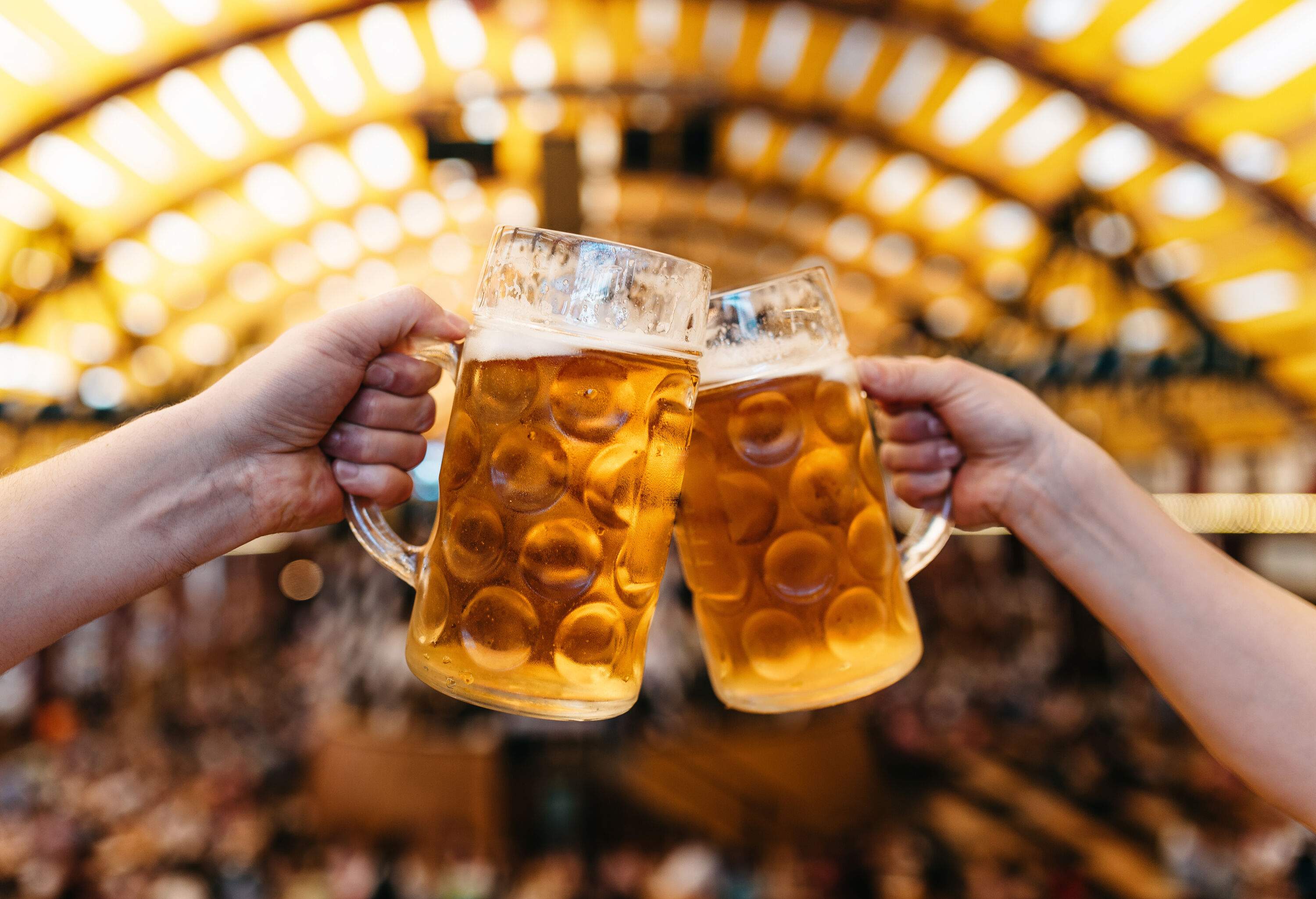 Two hands raise large mugs filled with beer against a bright bokeh background.