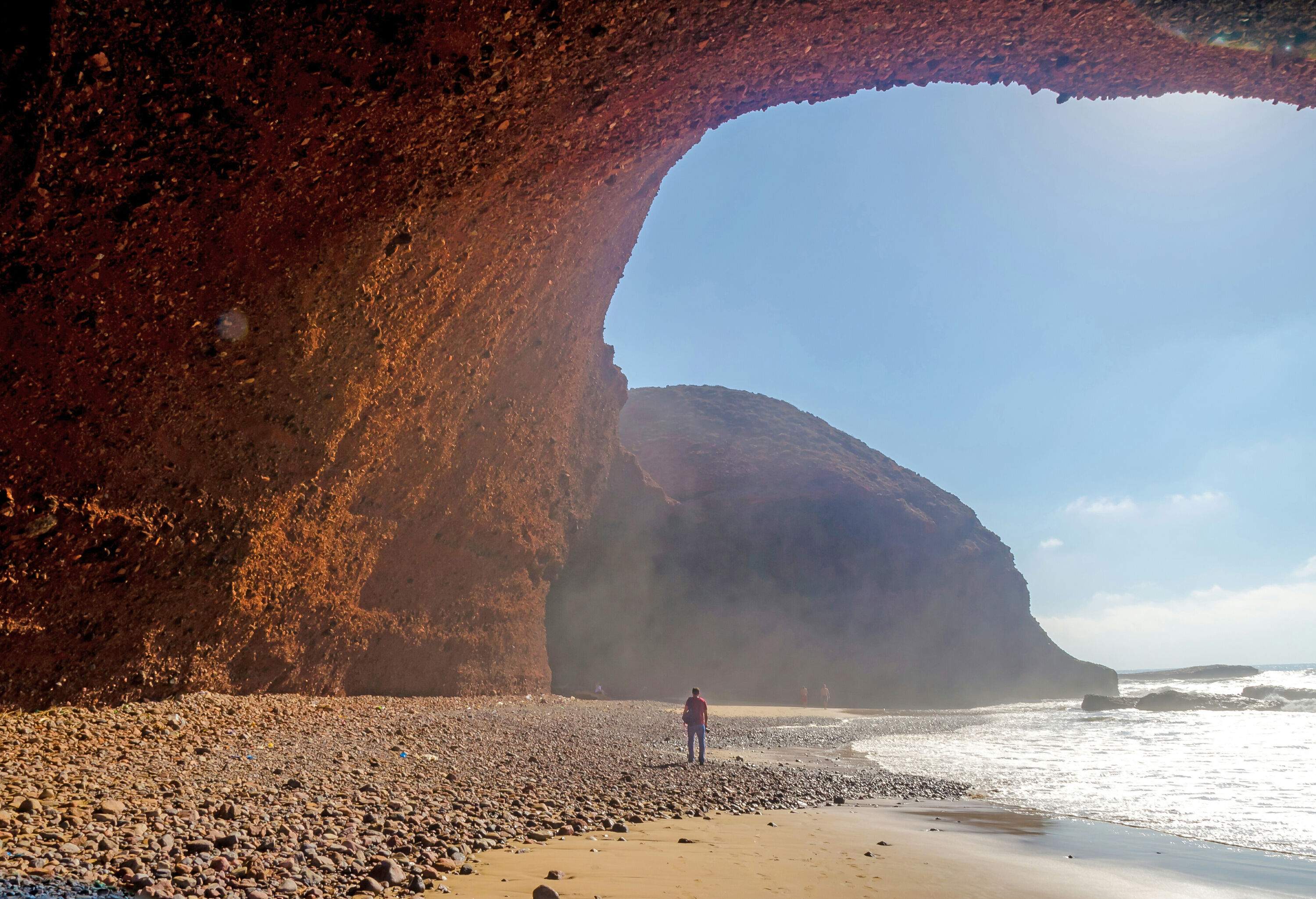 A person strolls across a pebbled beach under an arched rock formation.