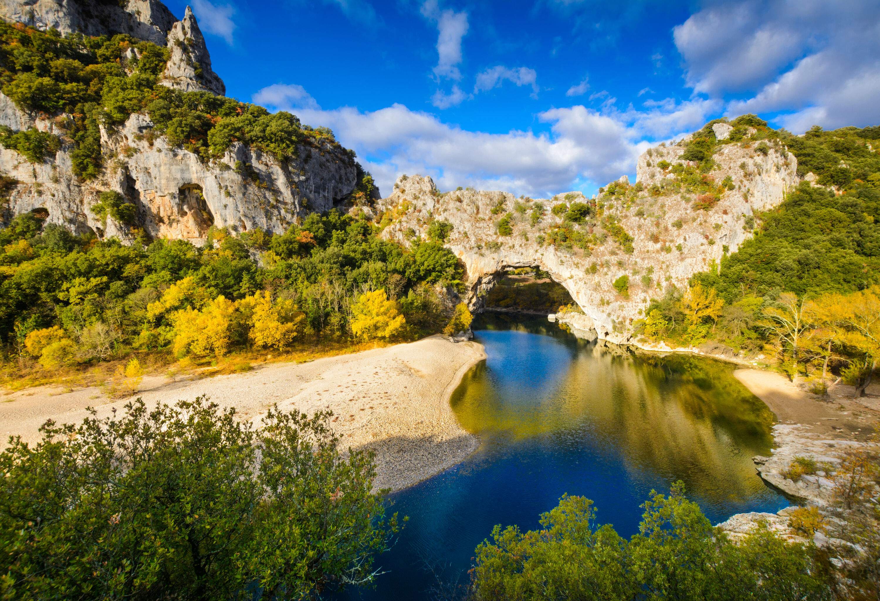 A natural sandstone arch bridge over a river surrounded by bushes.