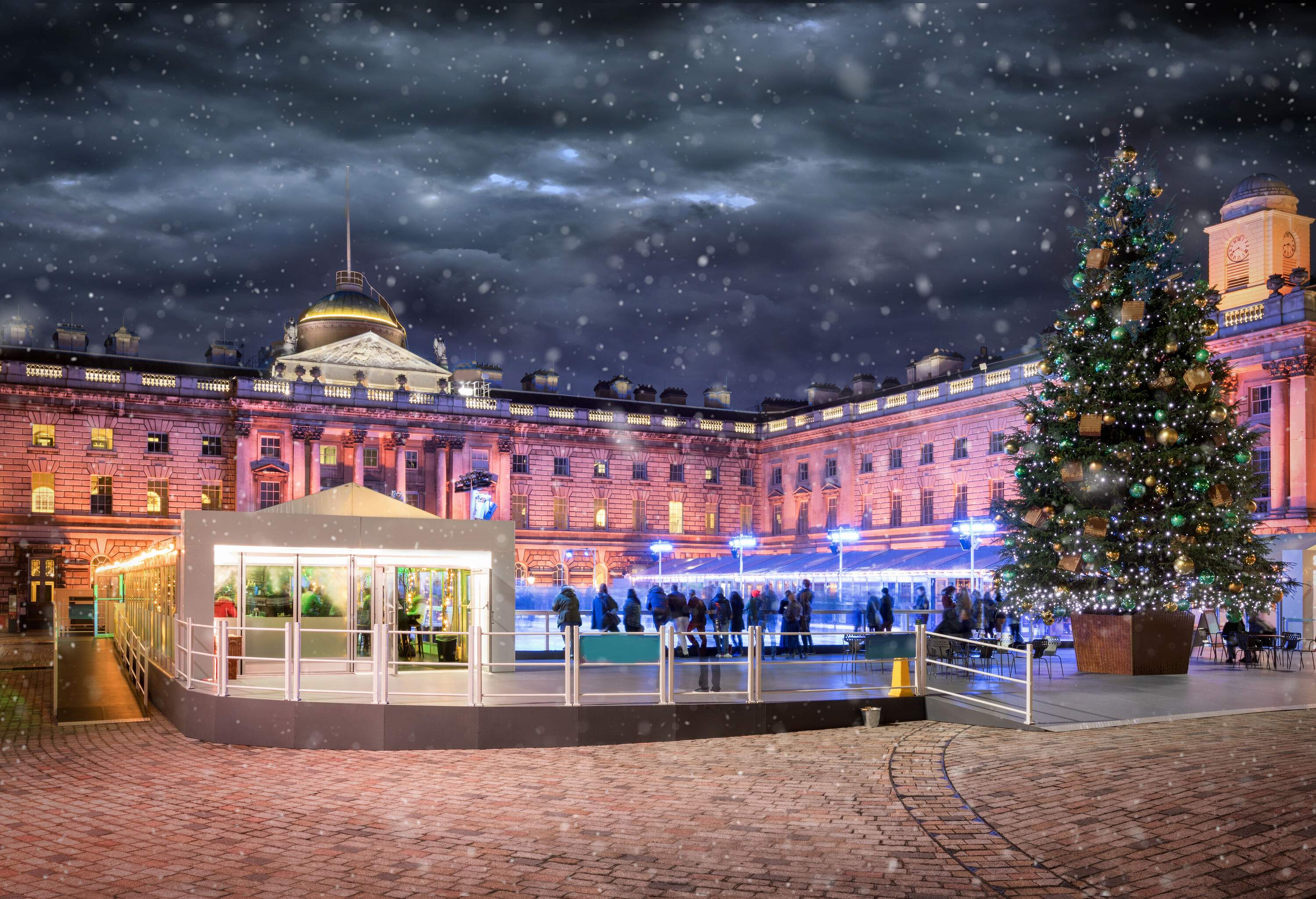 People at the ice skating rink in front of the illuminated Somerset House in London, England, decorated with a giant Christmas tree on a snowy winter night.