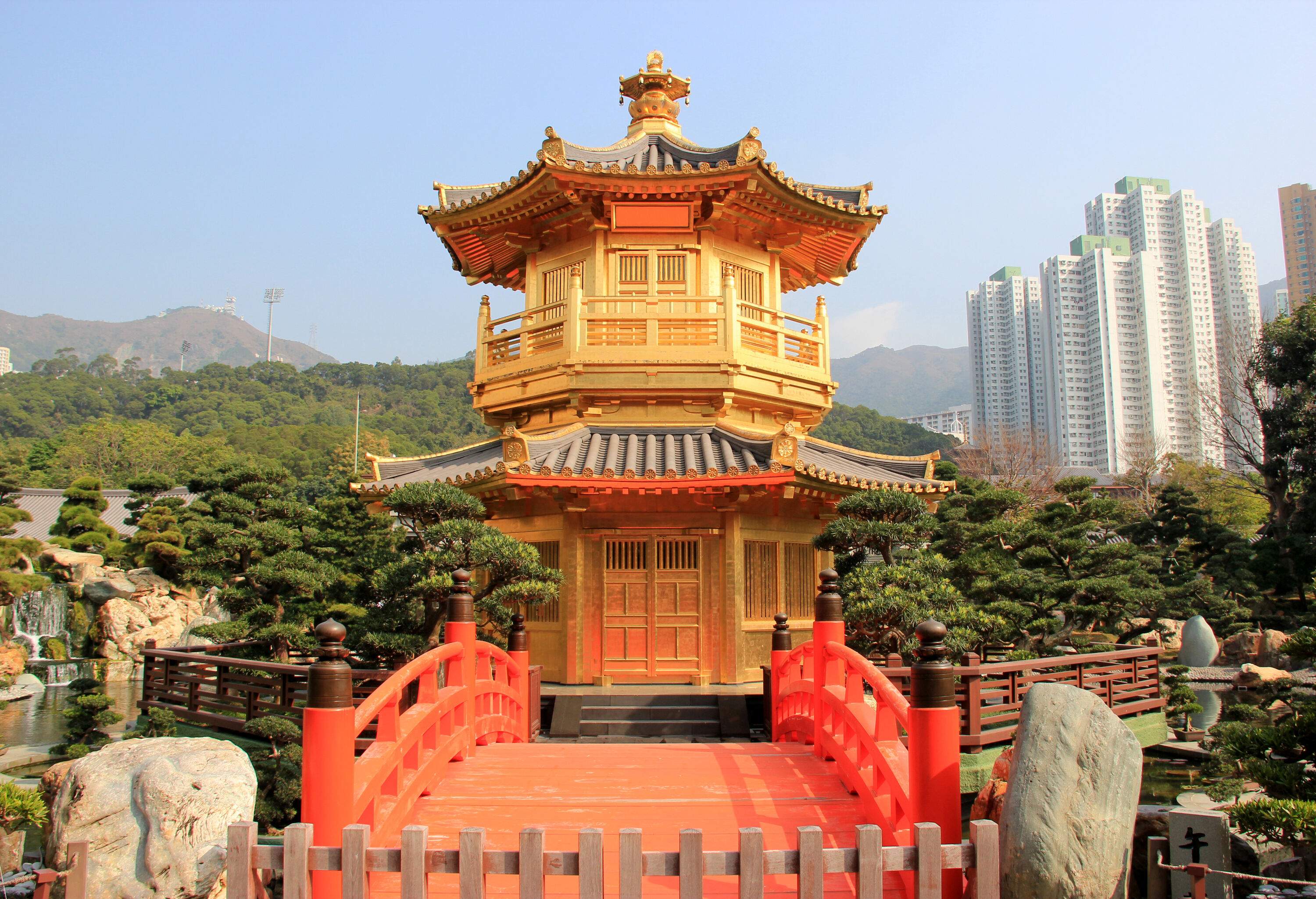 A two-storey golden pavilion accessed through a red bridge in a lush classical  Chinese garden.