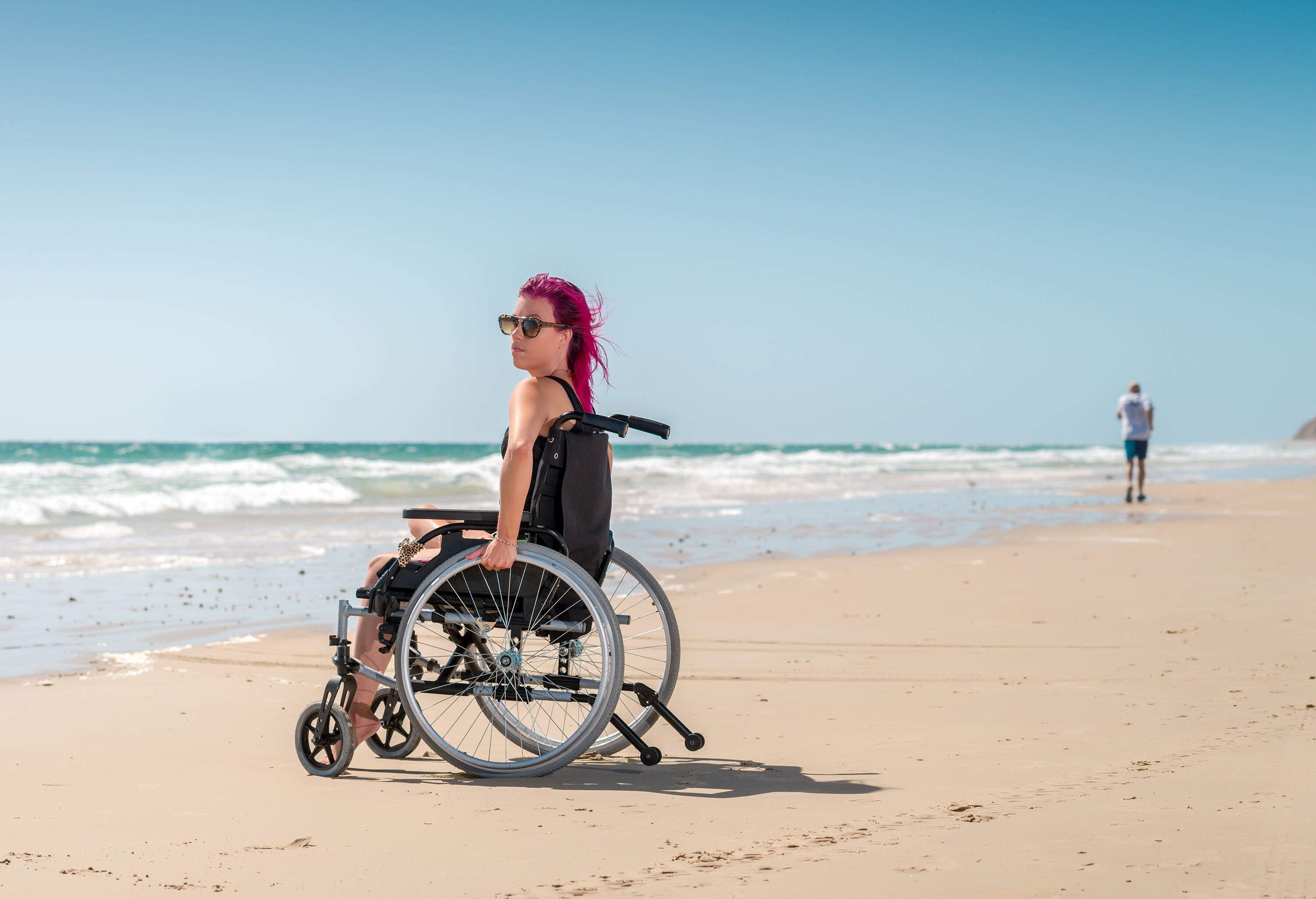 A woman with purple hair and wearing sunglass is sitting in a wheelchair on the shore of the beach.