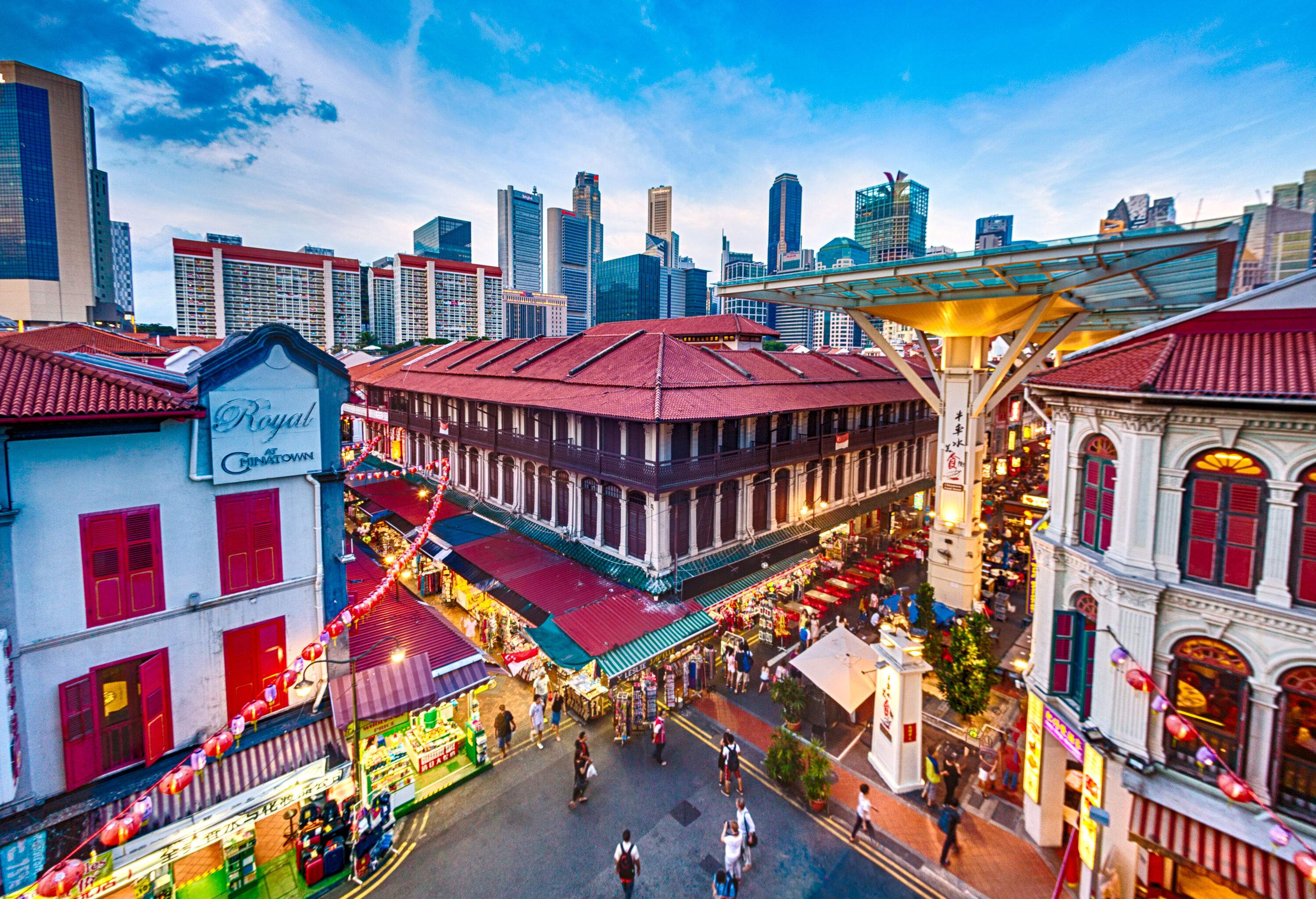 A commercial district with a crowded street lined with vendor stalls and restaurants with distant views of the city skyline.