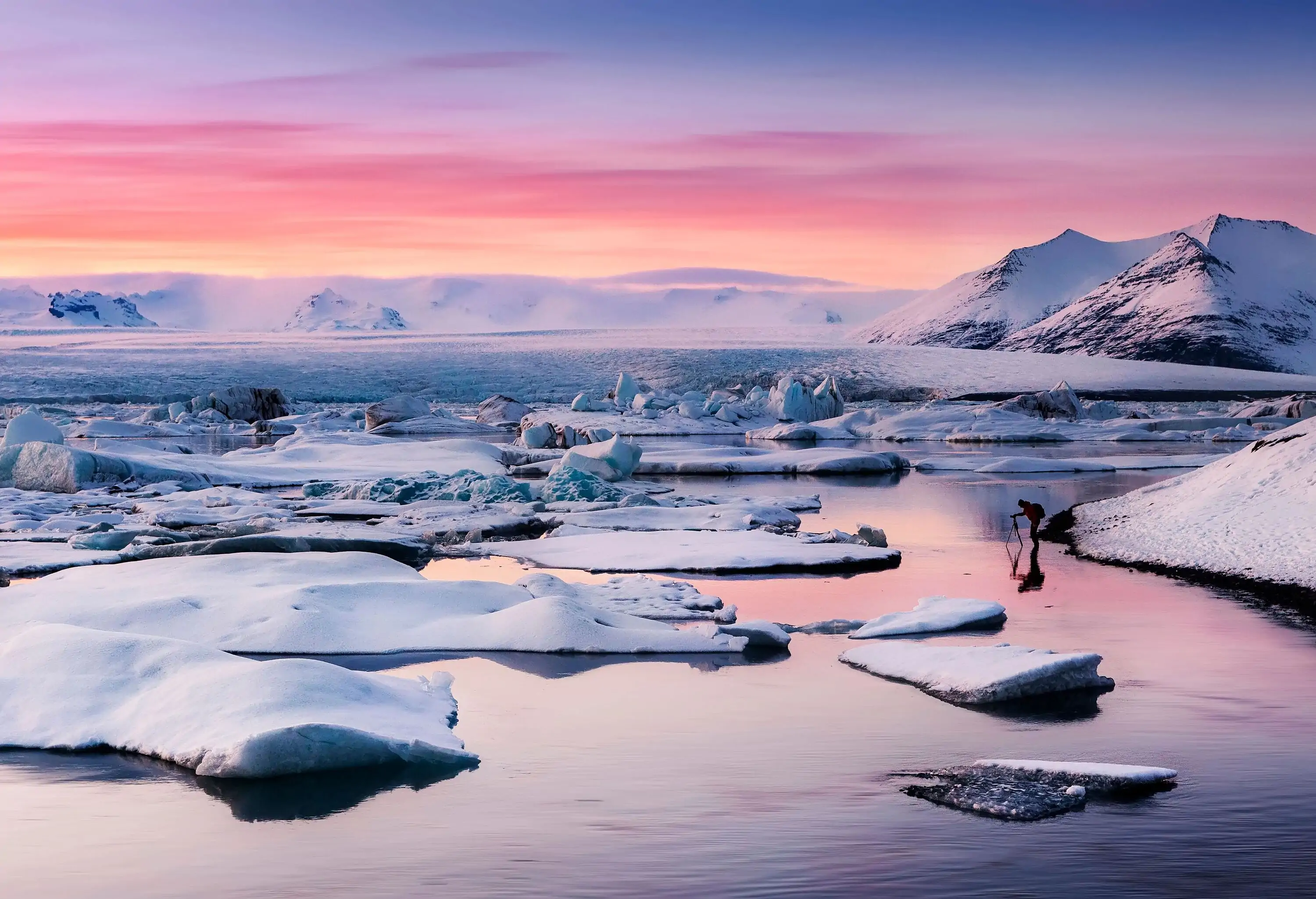 A picturesque lagoon with tranquil blue water and chunks of floating ice, nestled amidst snow-covered hills in the distance.