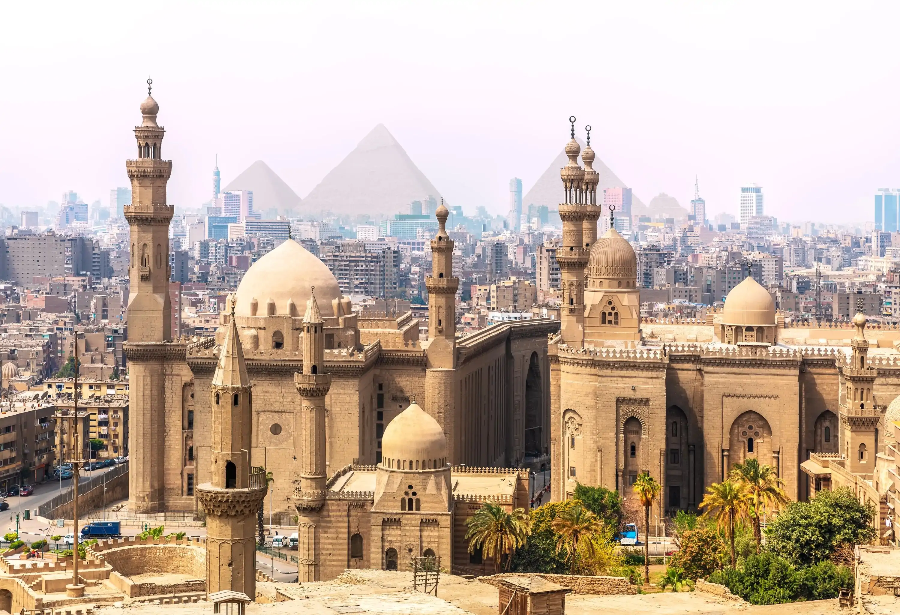 A vast mosque with unique architectural features and towering minarets, in the centre of an old town with traditional architecture buildings and pyramids in the background.