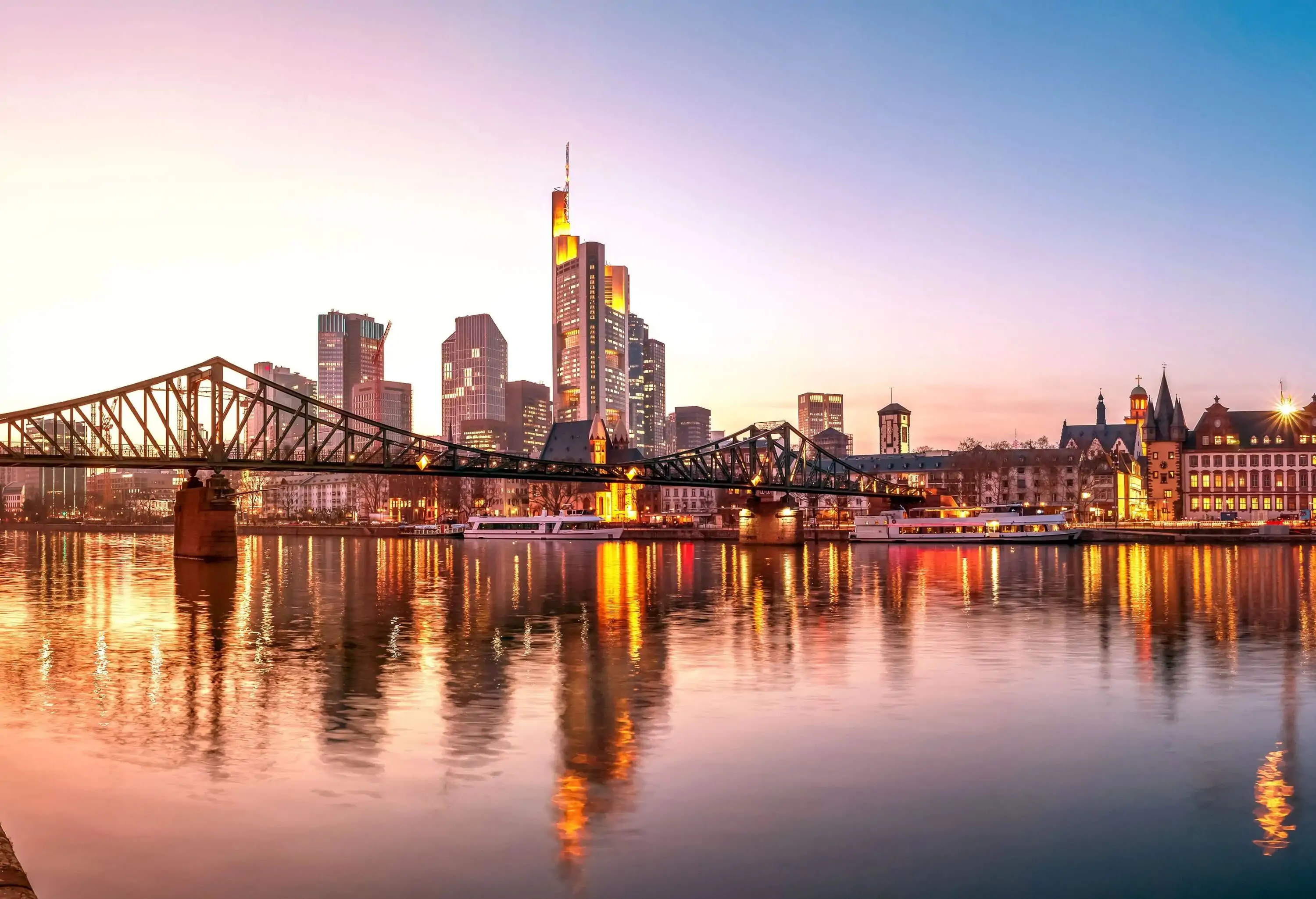 A long steel modern bridge over the river against the illuminated riverside cityscape with a mixture of old and modern buildings with skyscrapers.