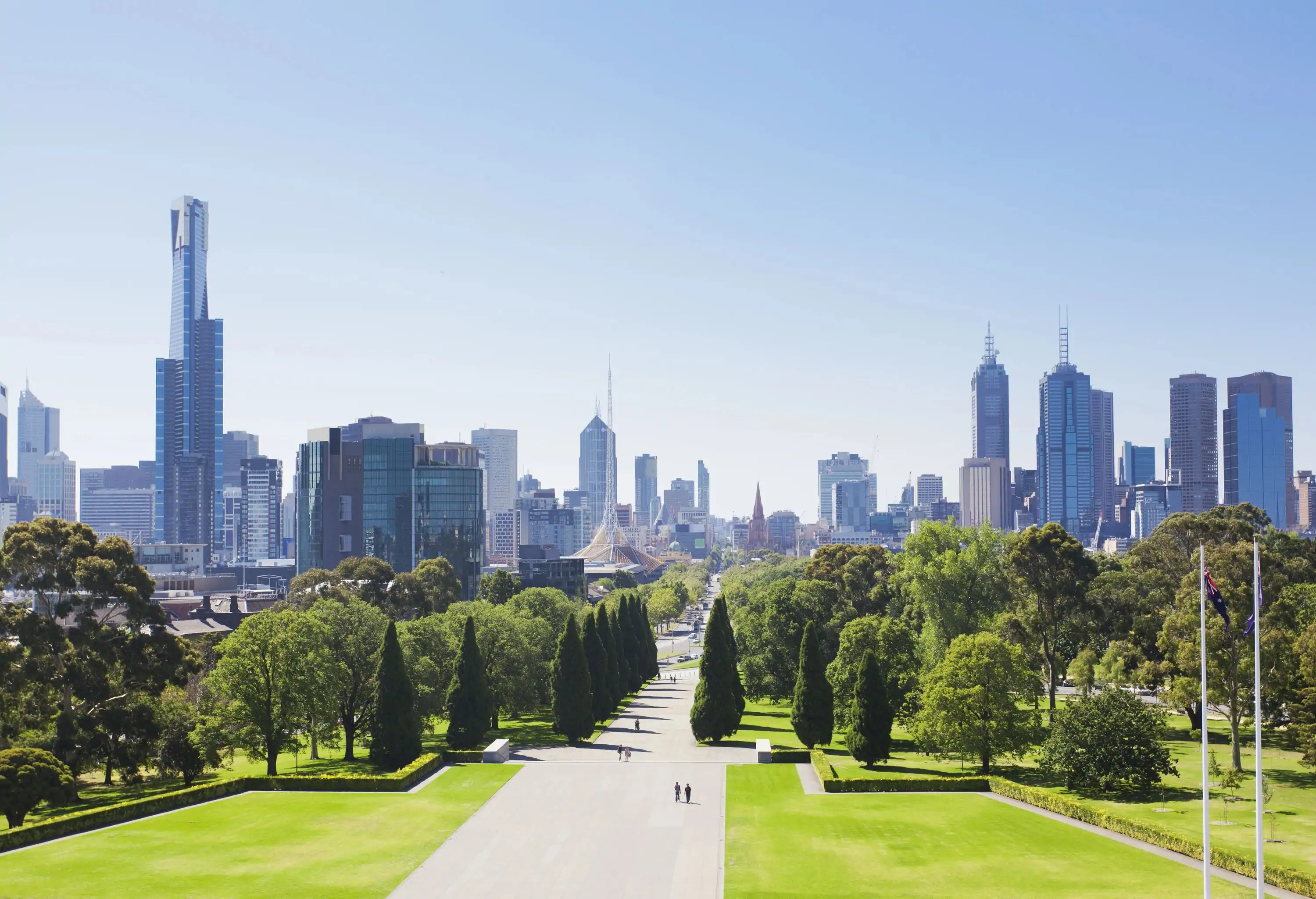 Several people walk on the broad paved walkway in a lush green park surrounded by tall buildings.