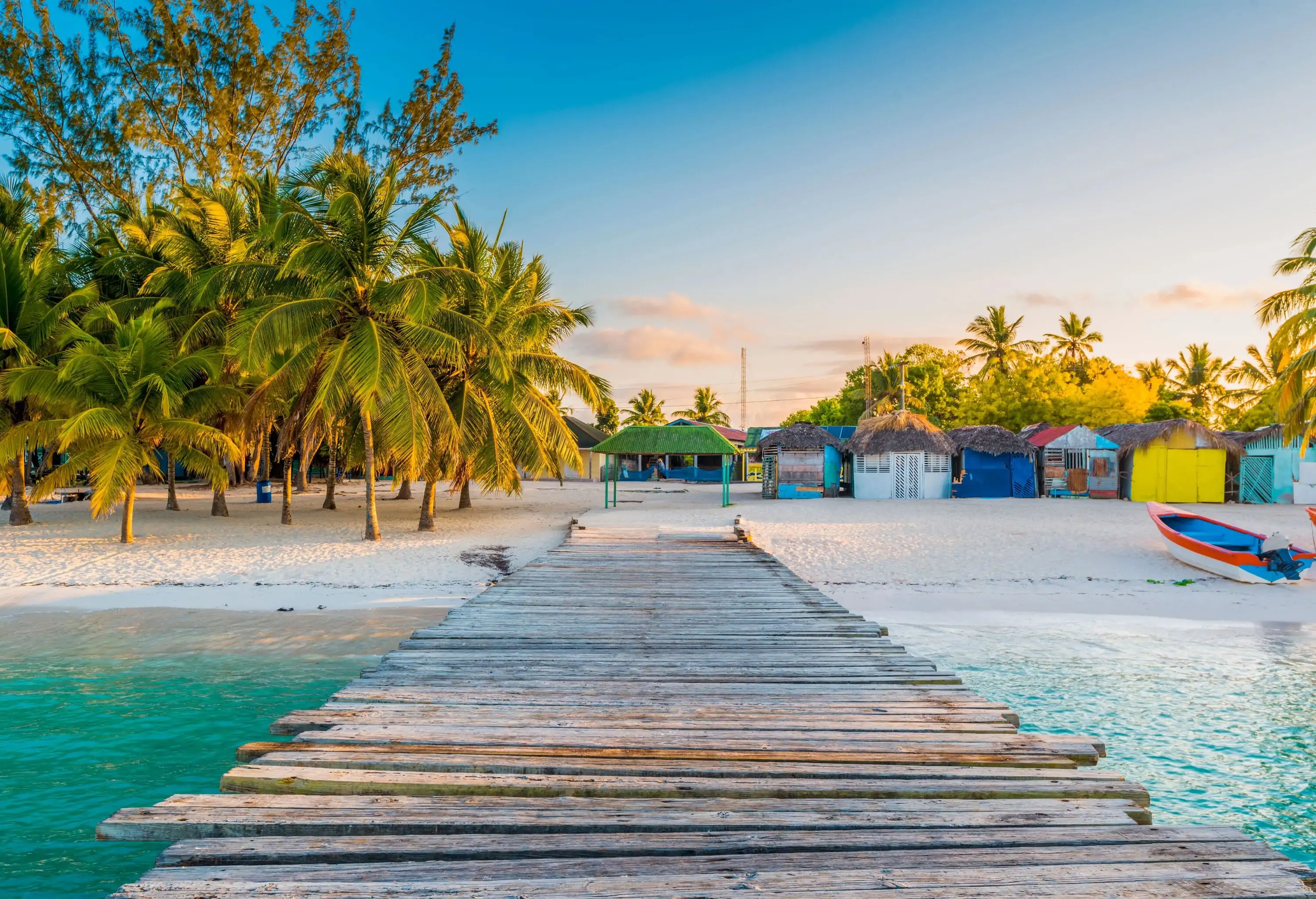 A boardwalk stretching over the turquoise sea to a sandy white beach dotted with coconut trees and colourful cabins.