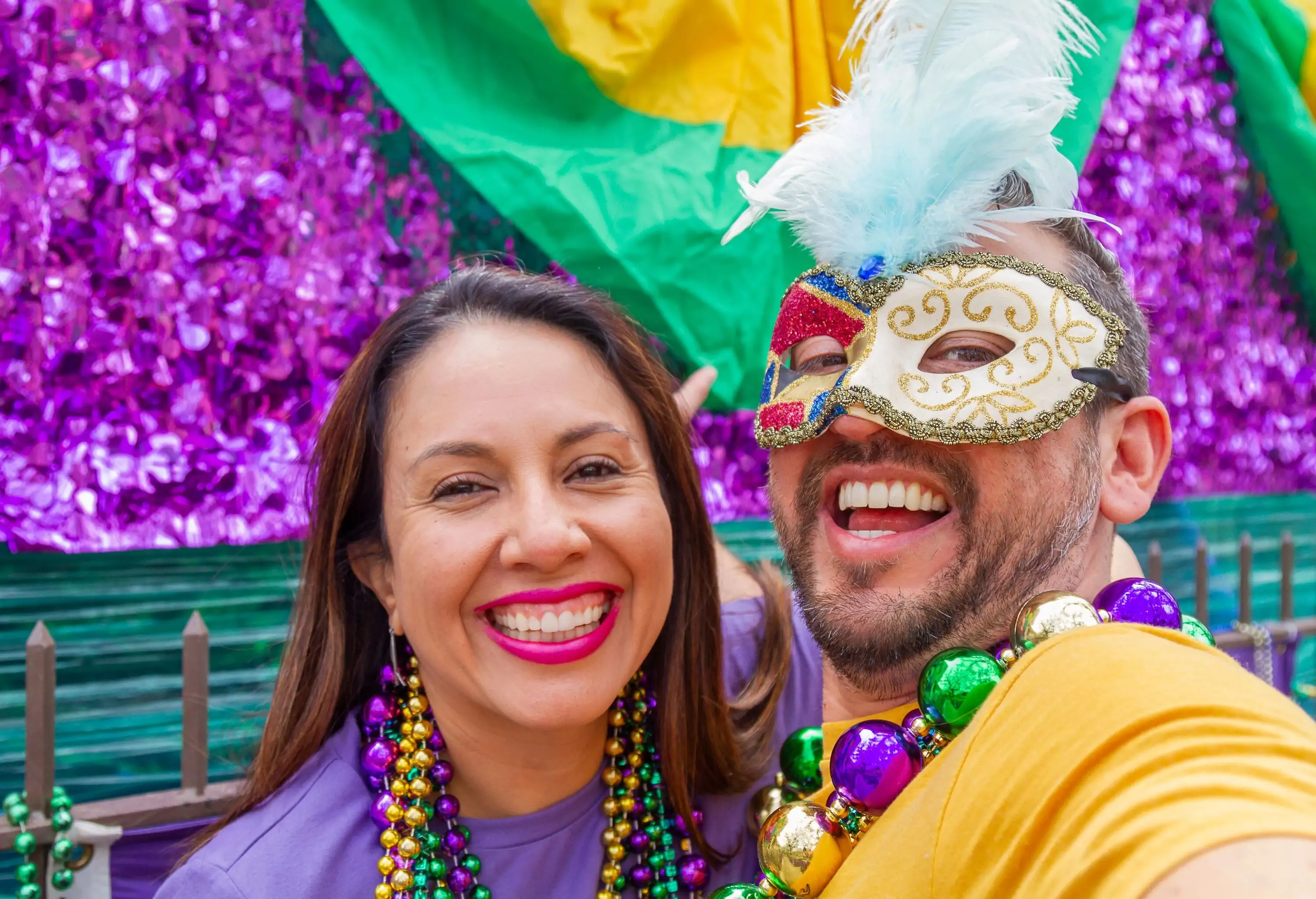 A happy couple smiles as they wear vibrant shirts, colourful bead necklaces, and carnival mask.