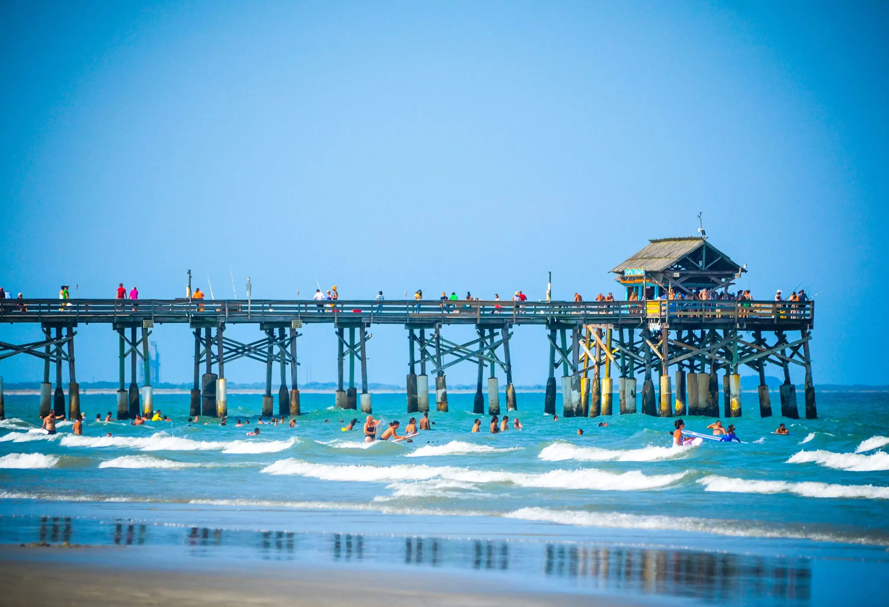 Tourists stroll on a long pier as they watch a group of people swimming on a beach with mild waves.