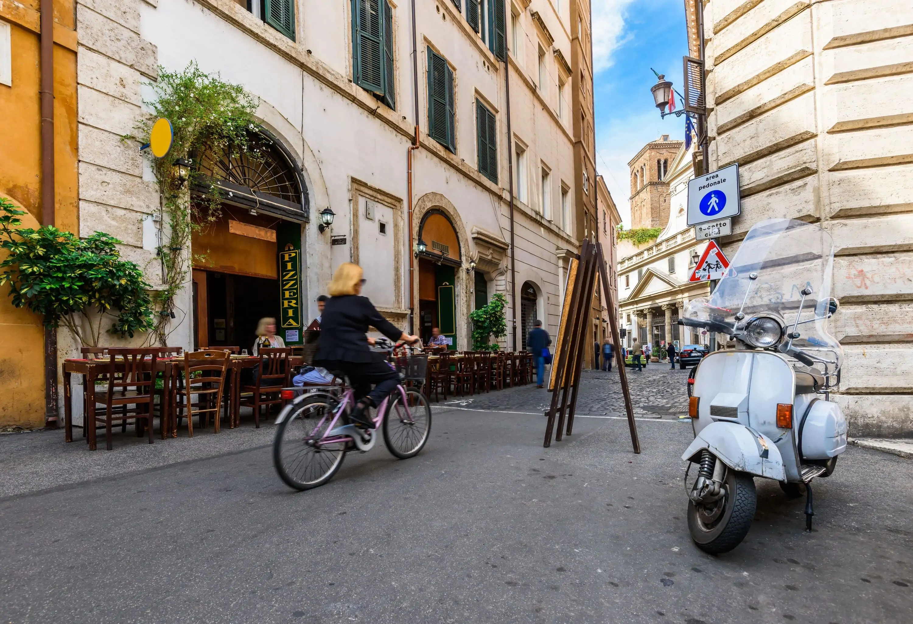 A blonde woman riding her bike through a tiny street surrounded with outdoor cafes.