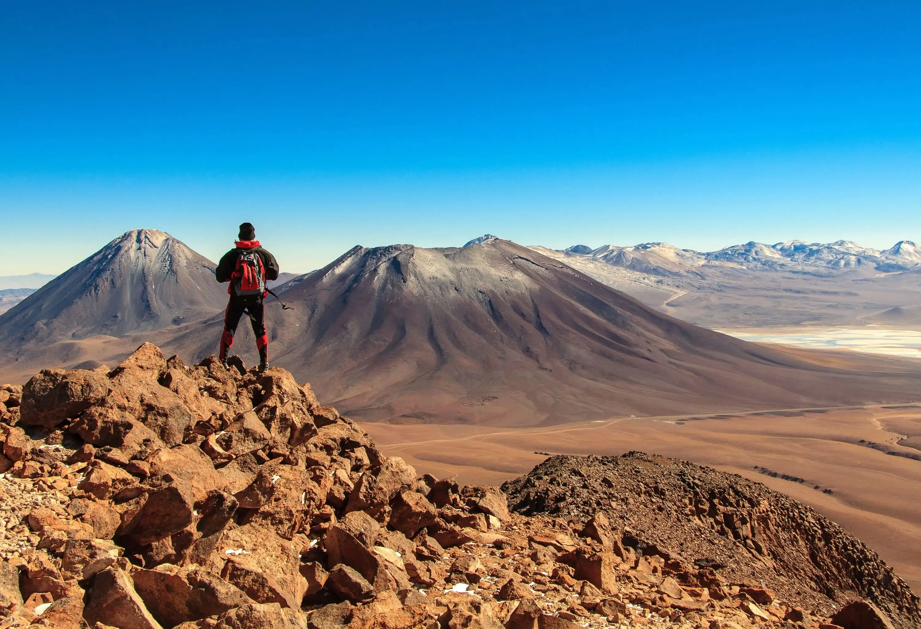 A hiker on a rocky hilltop overlooking a dormant volcano under the scenic clear blue sky.