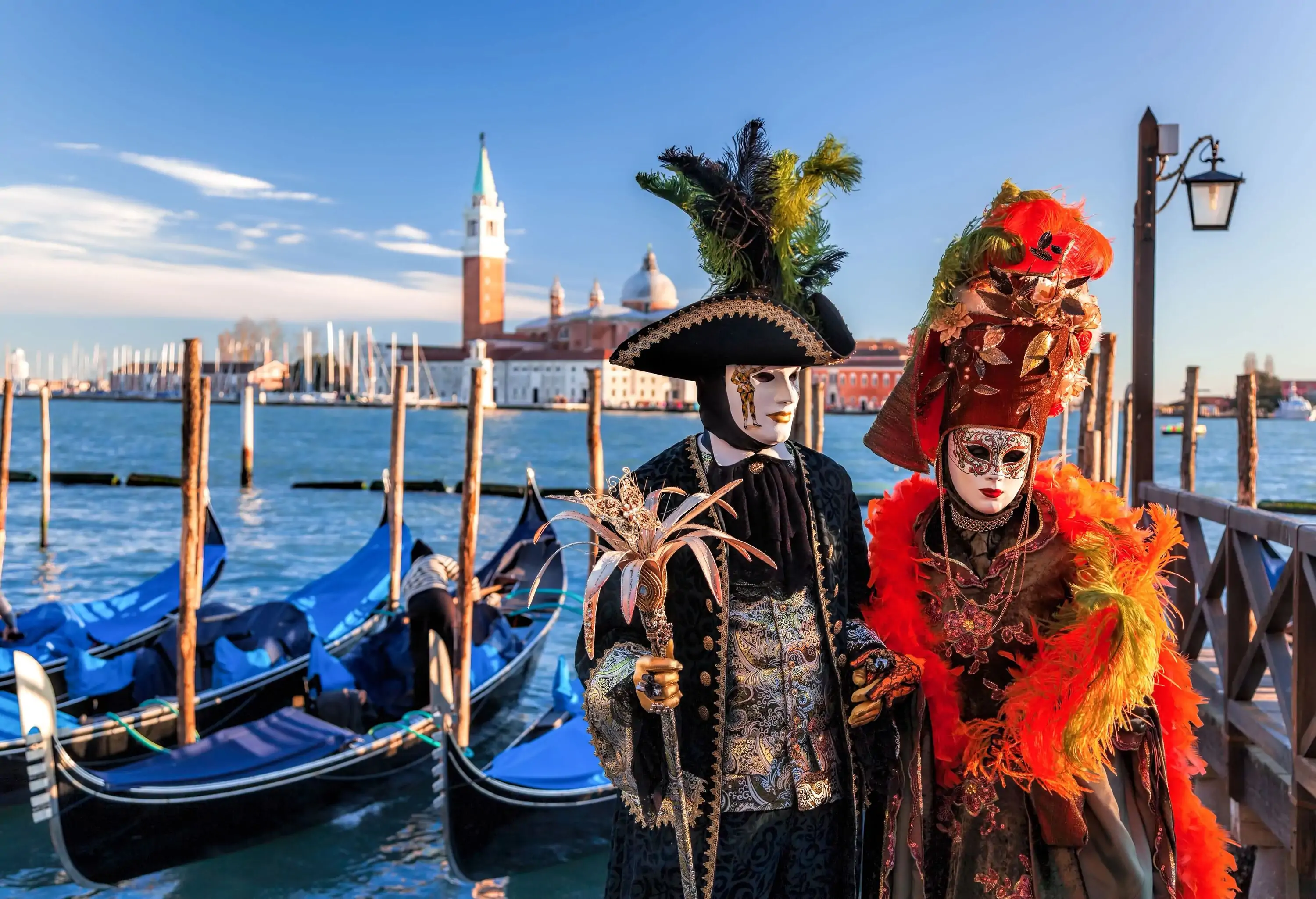 Two person dressed in elegant festival costumes with black robe, orange feathers, white carnival masks, and ornamental hats.