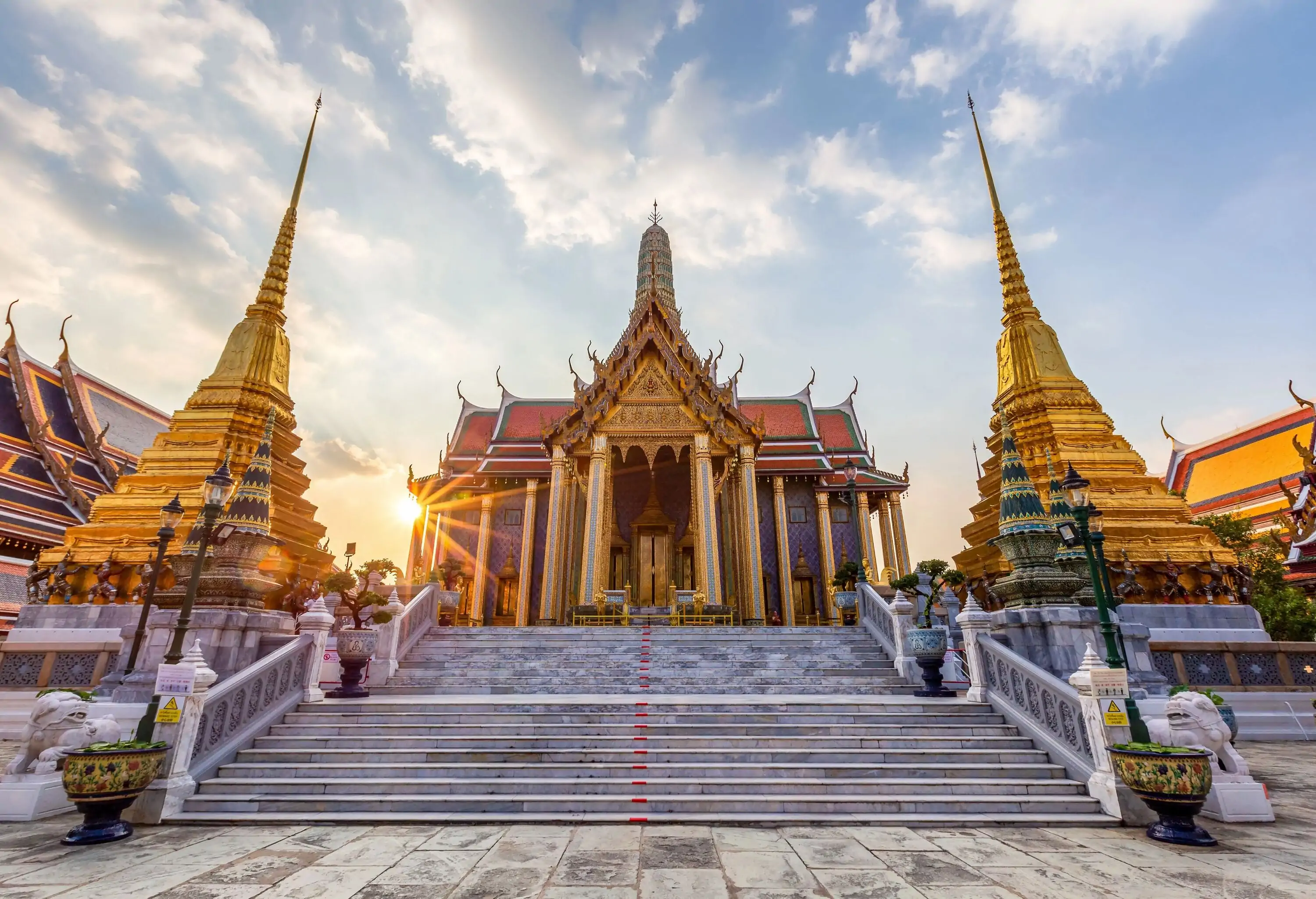 The Temple of the Emerald Buddha is a red-roofed temple with beautiful golden carvings on the gable, supported by gold-plated pillars, and positioned between two golden bell-shaped stupas.