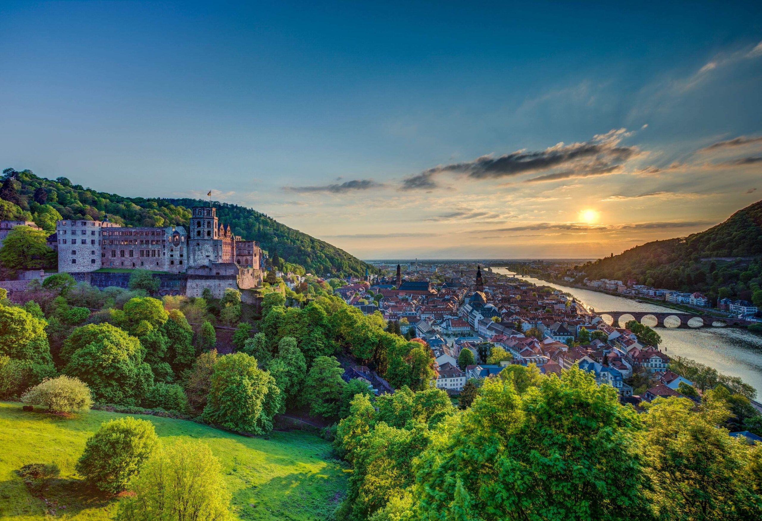 A hilltop castle with a view of a riverside town and the sun setting in the horizon.