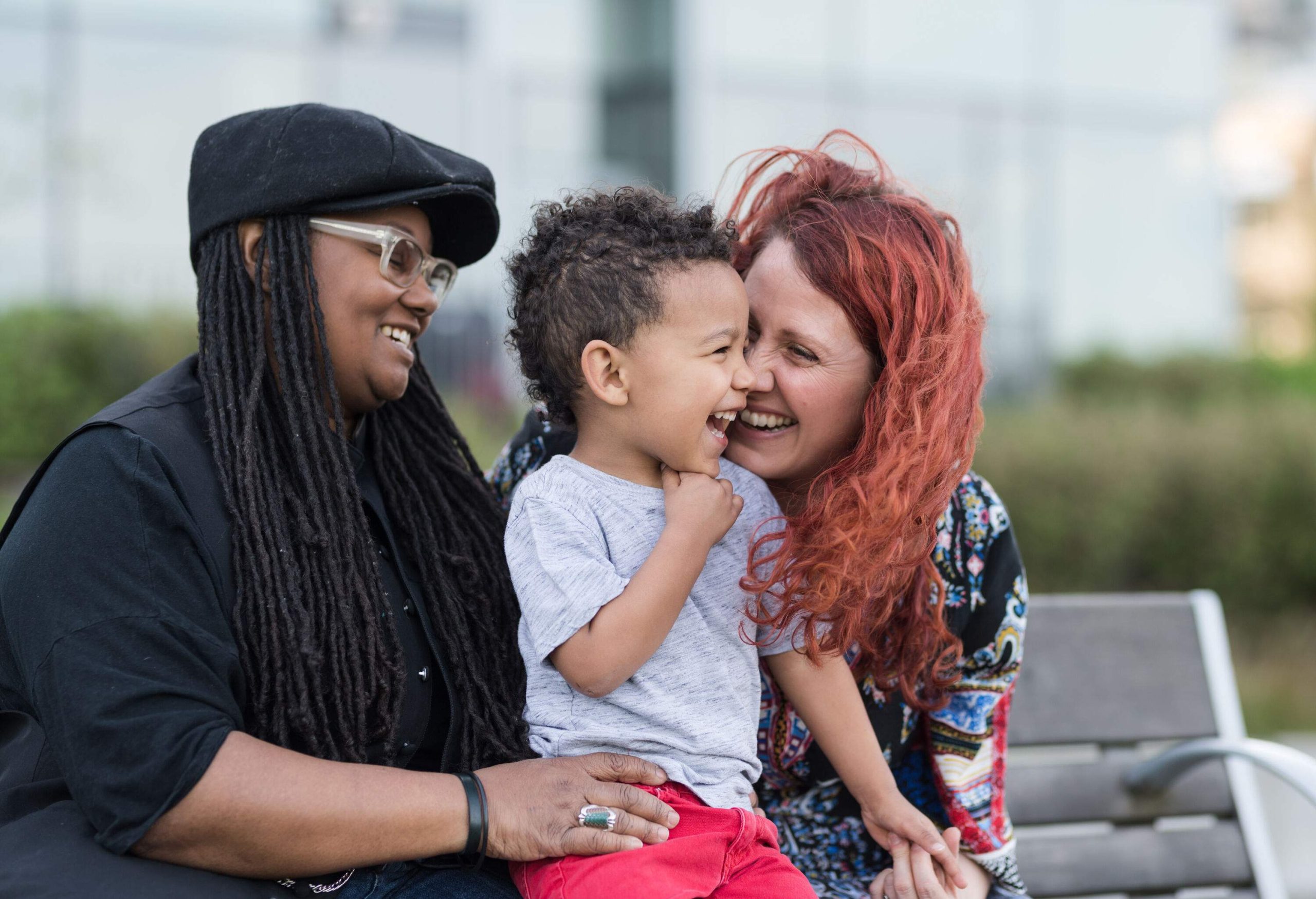 A happy couple sits on a park bench with their young son sitting on their laps.