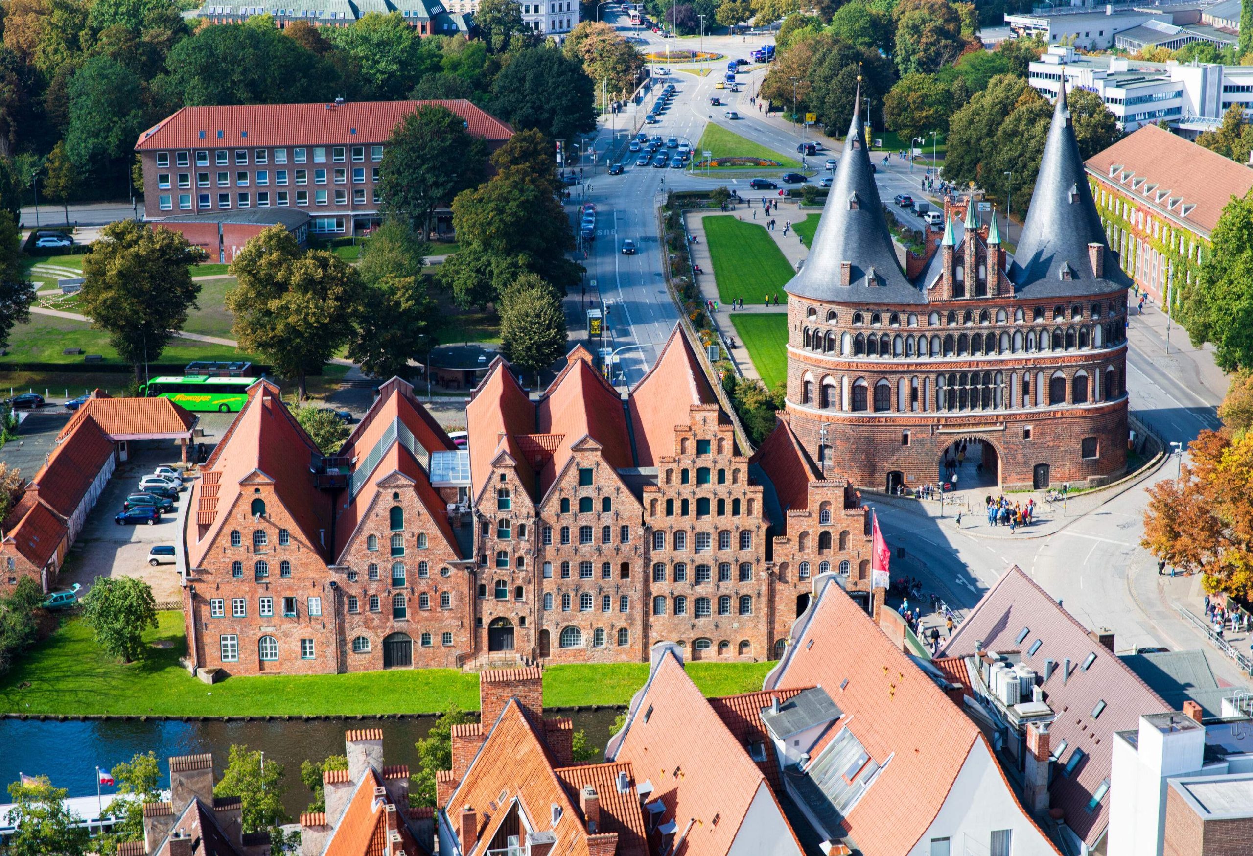 The Holsten Gate, a gatehouse with linked towers on both sides with pointed roofs, is adjacent to a plaza surrounded by buildings.