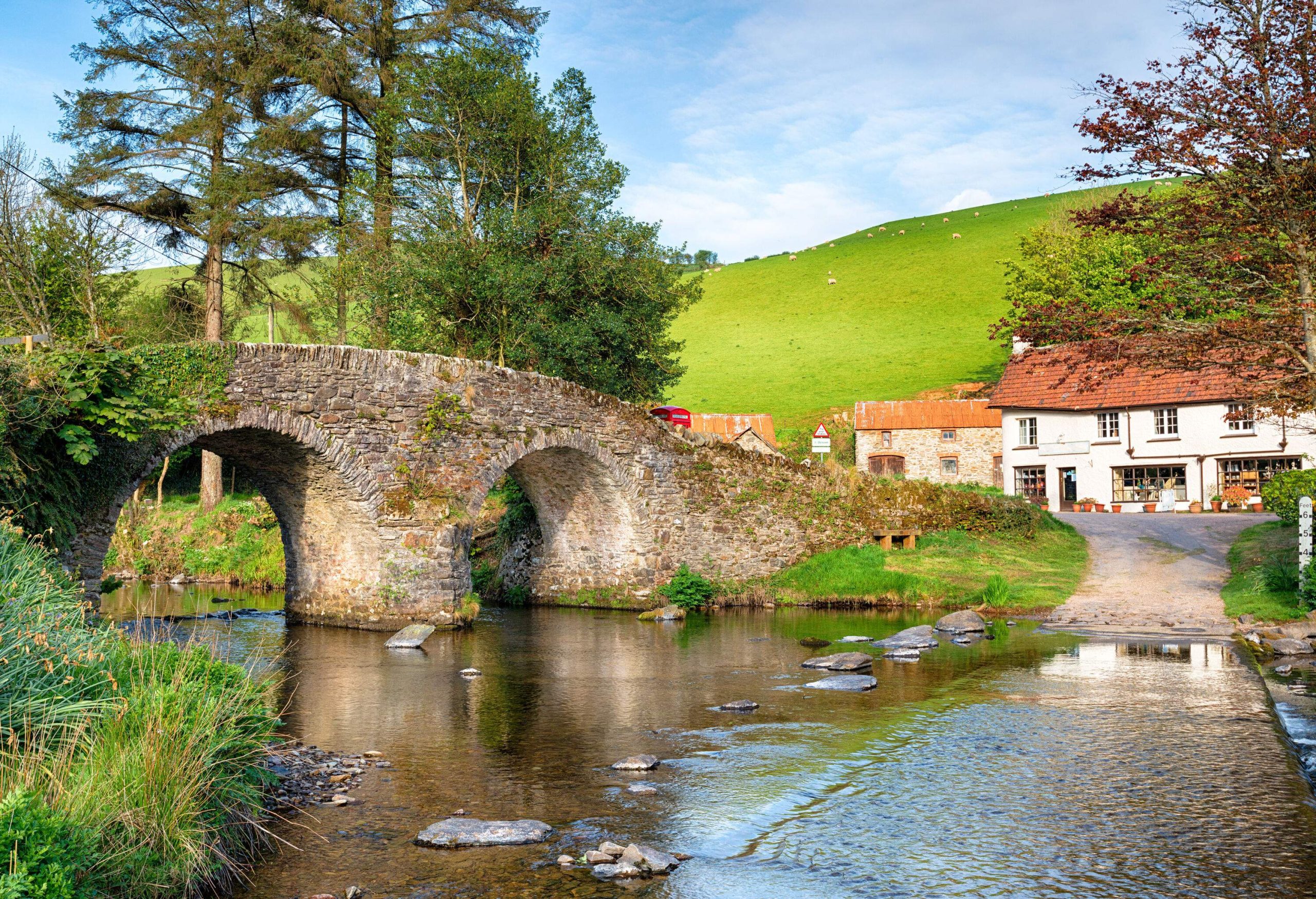 A stone bridge with arches above the shallow river and houses beneath a lush hill.