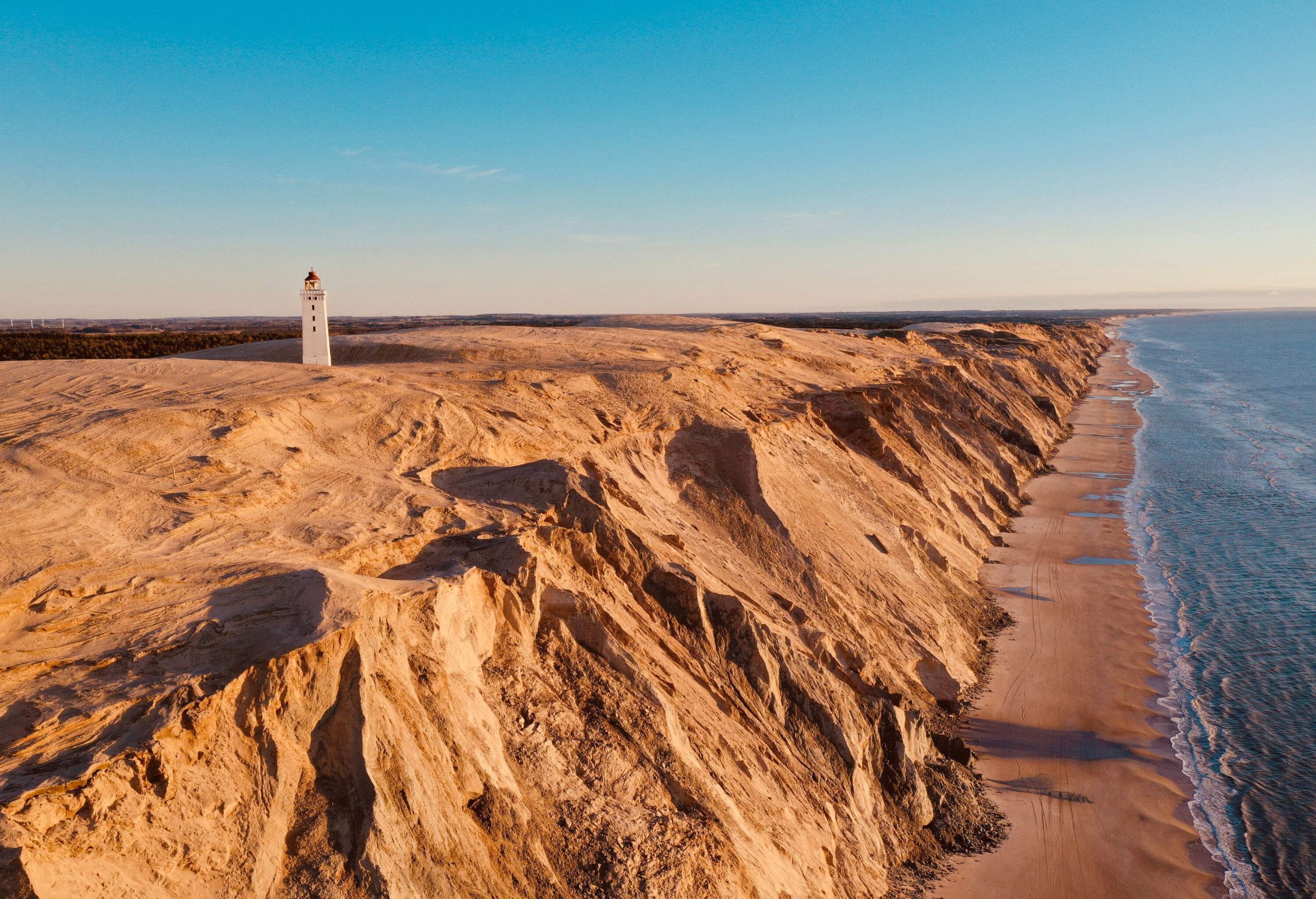 A white tower topped with a red lantern lighthouse encroached on the dunes surrounded by the sea under the blue sky.