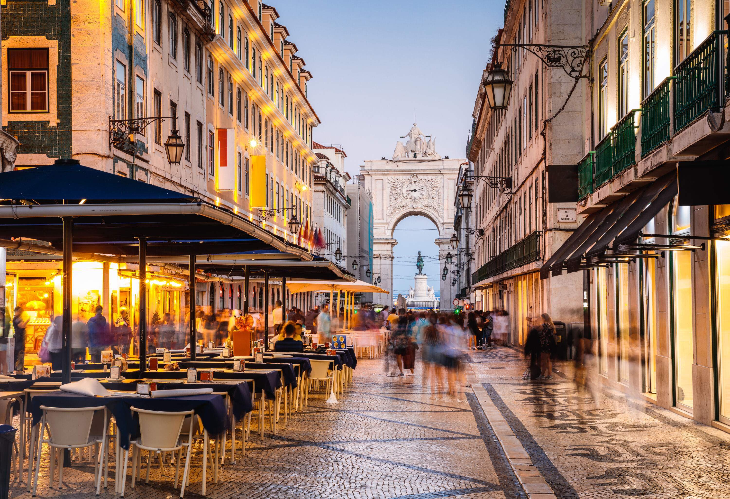 Tourists pass through the cobbled road with outdoor dining between buildings towards an archway.