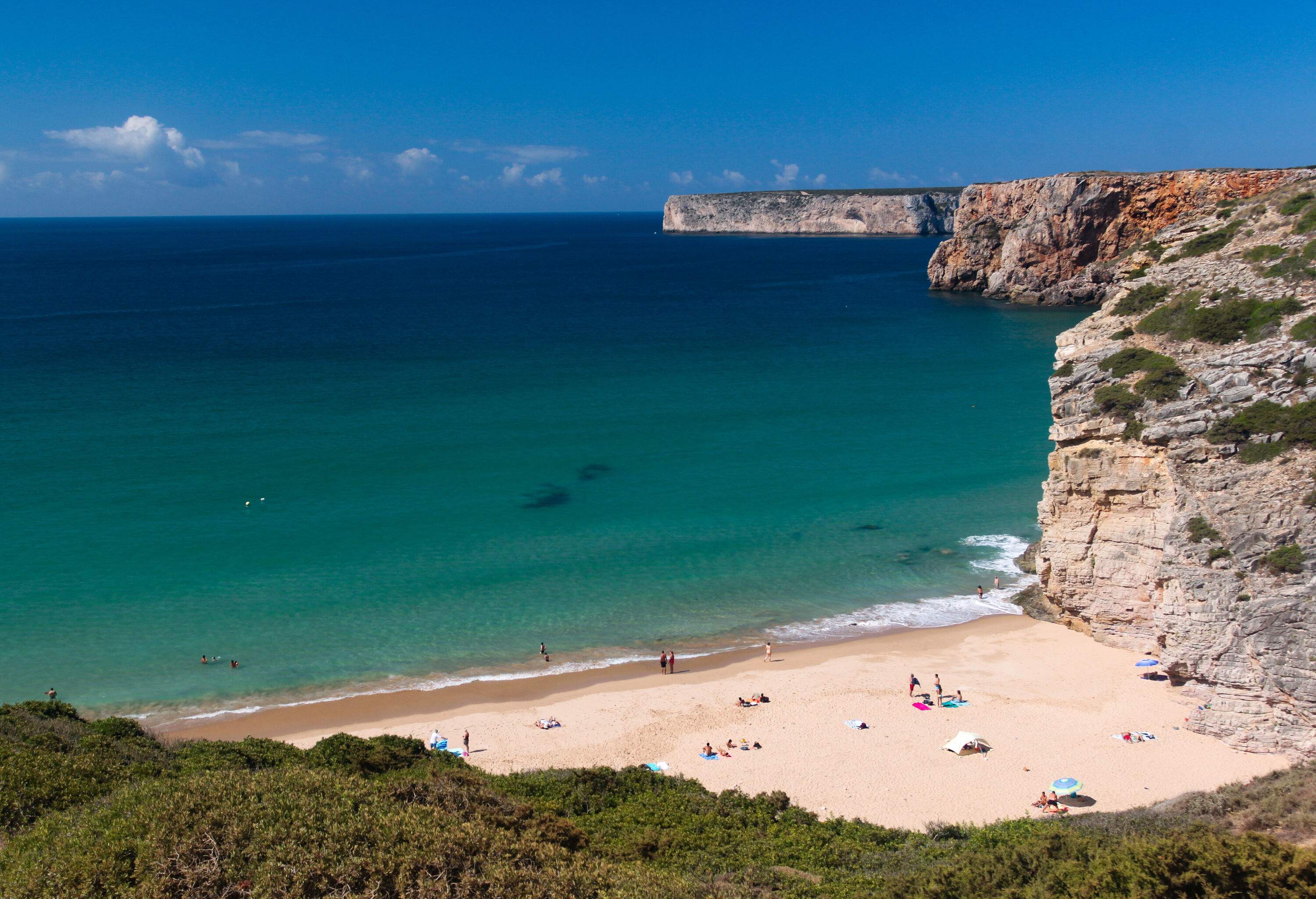 A sandy shore stretches alongside the tranquil blue sea, encompassed by expansive cliffs under the clear blue sky.