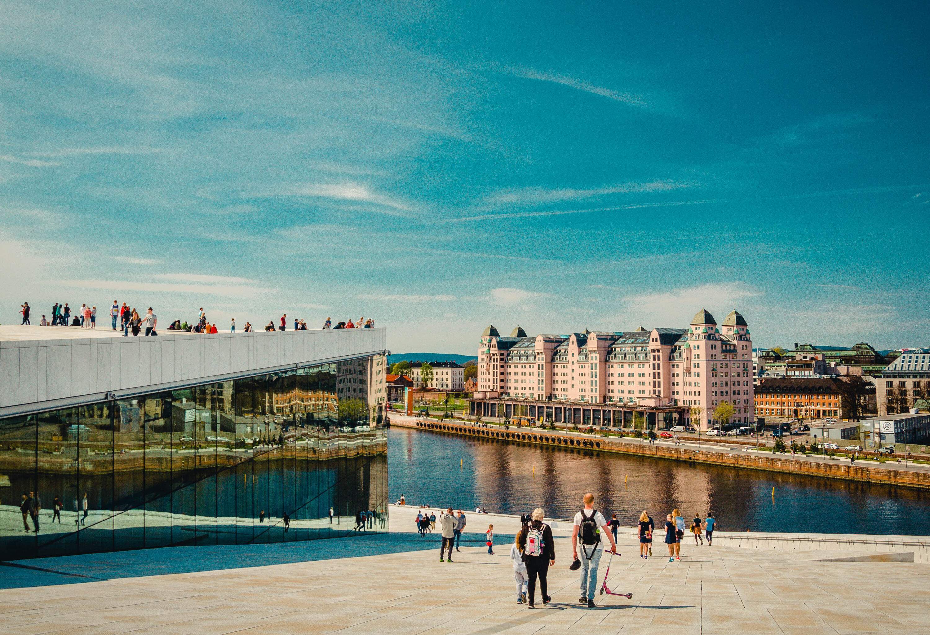 A group of people leisurely stroll along a sloped boardwalk that leads to the river, passing by a modern building with glass walls, all against the backdrop of other buildings on the opposite side of the river.