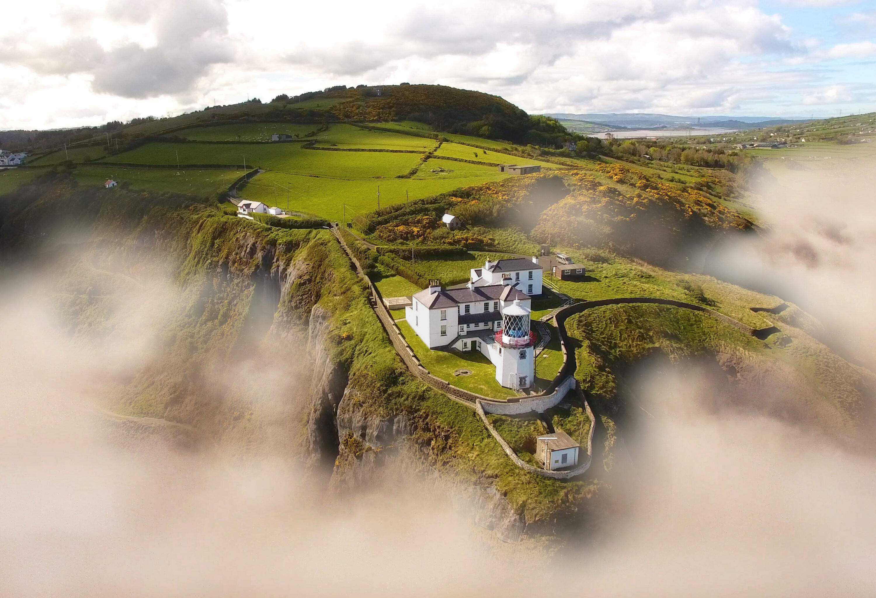 A lighthouse located on the edge of a cliff surrounded by lush grasslands, with fog clinging along the ridges.