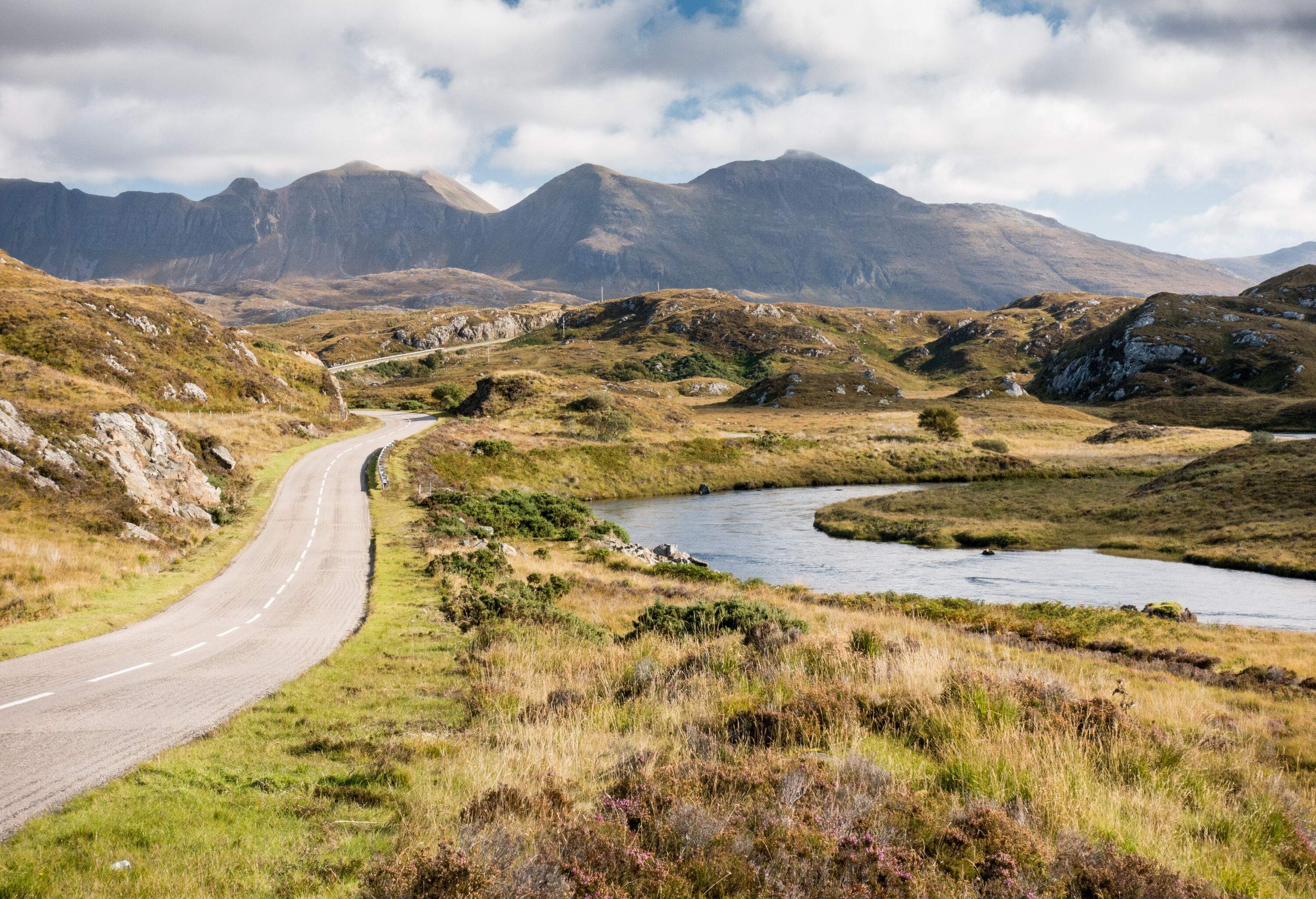 A road runs along a river and low-lying hills with distant views of a mountain range.