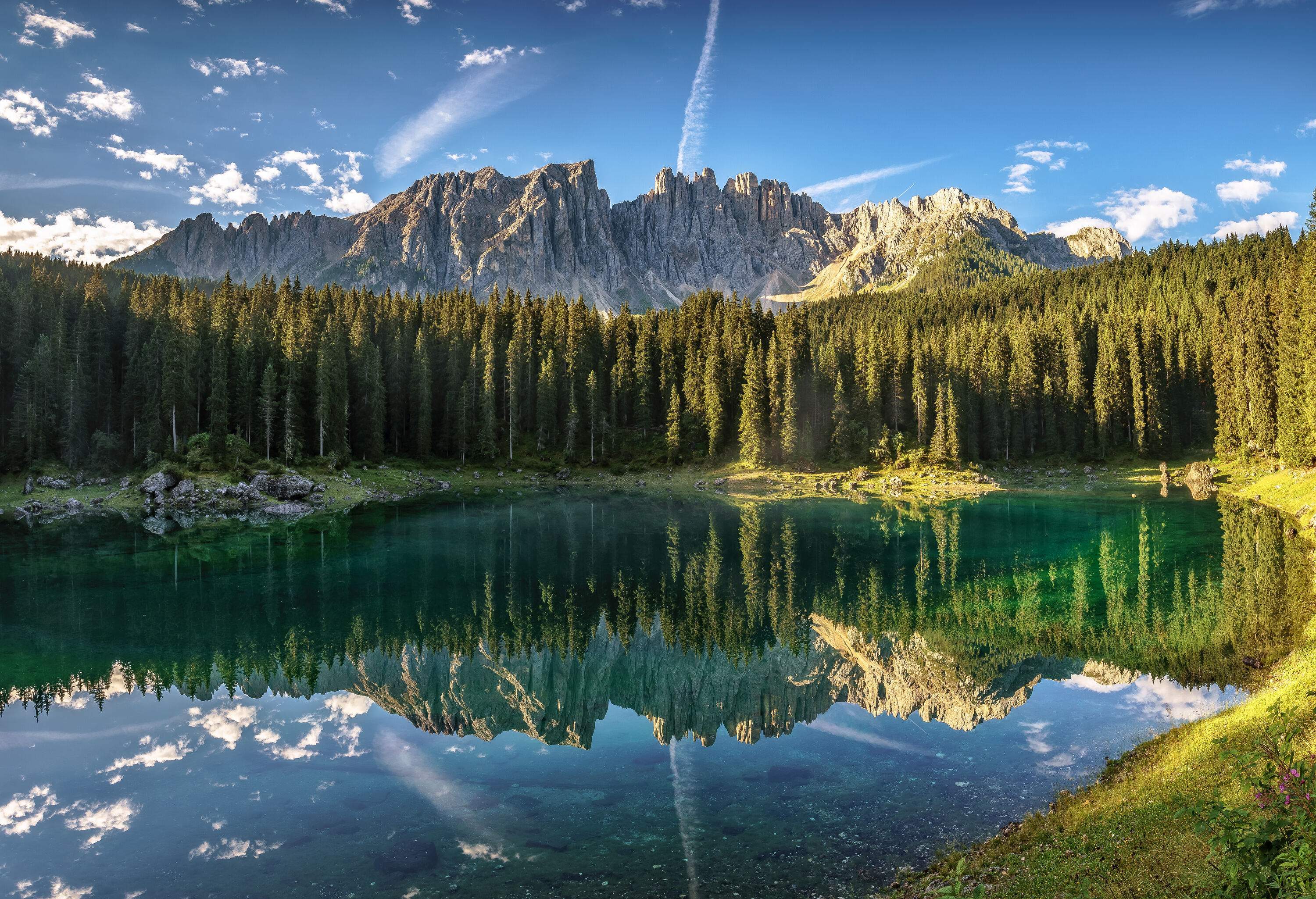 A picturesque lake with crystal-clear water mirrors the surrounding trees and the mountain range in the background under a blue sky.