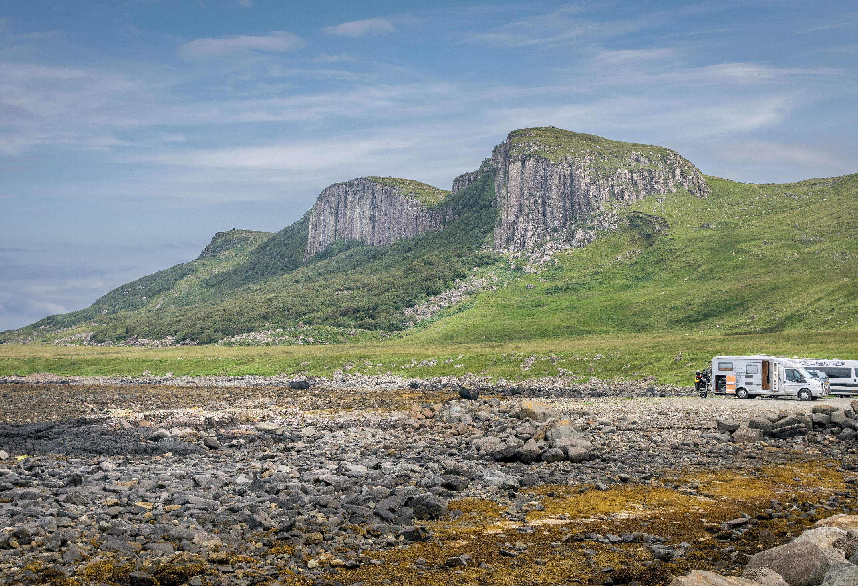 Vehicles parked on a rocky landscape beneath the mountains covered in a green meadow.