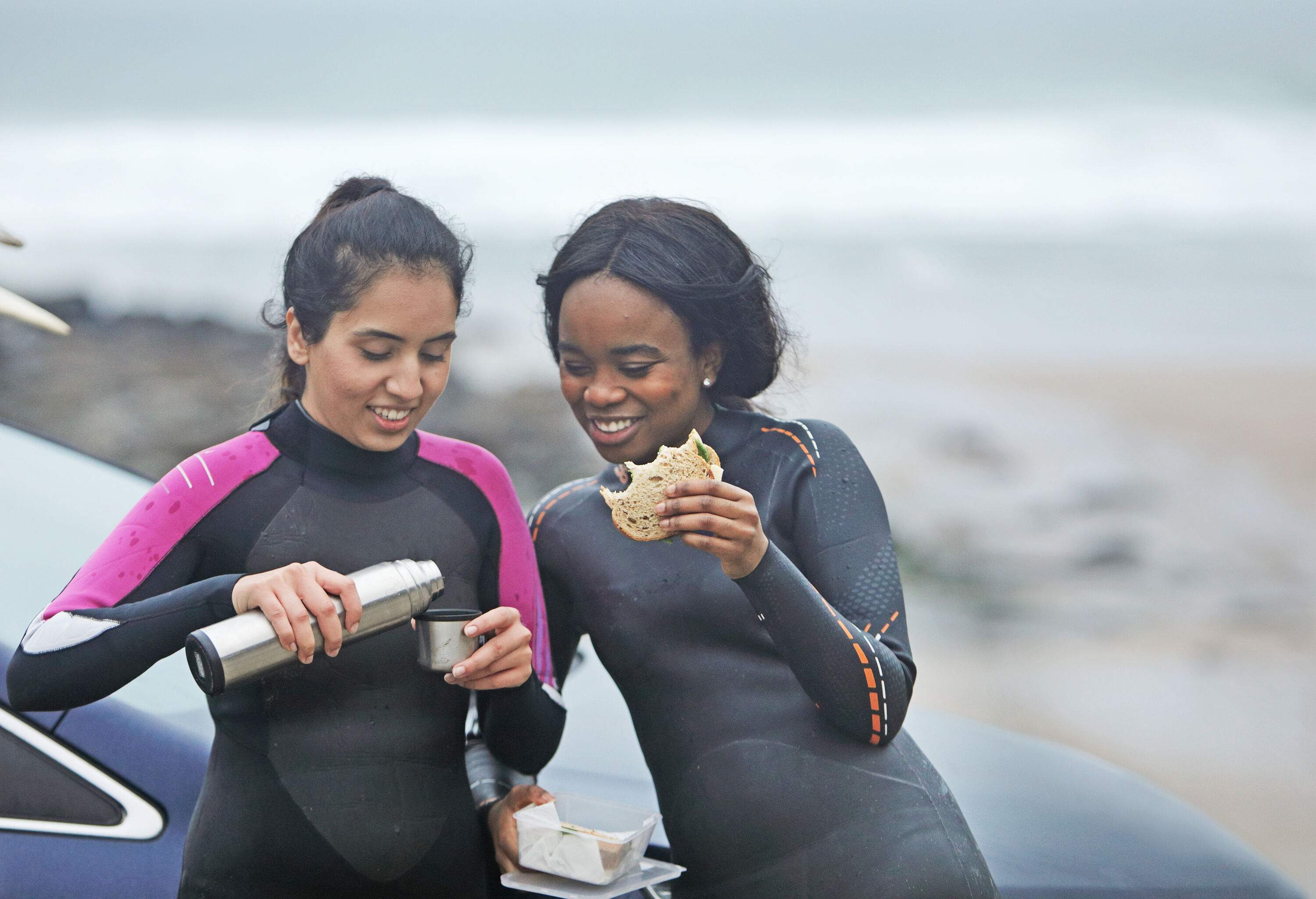 A couple of friends in wetsuits enjoying a drink and a sandwich by their car.