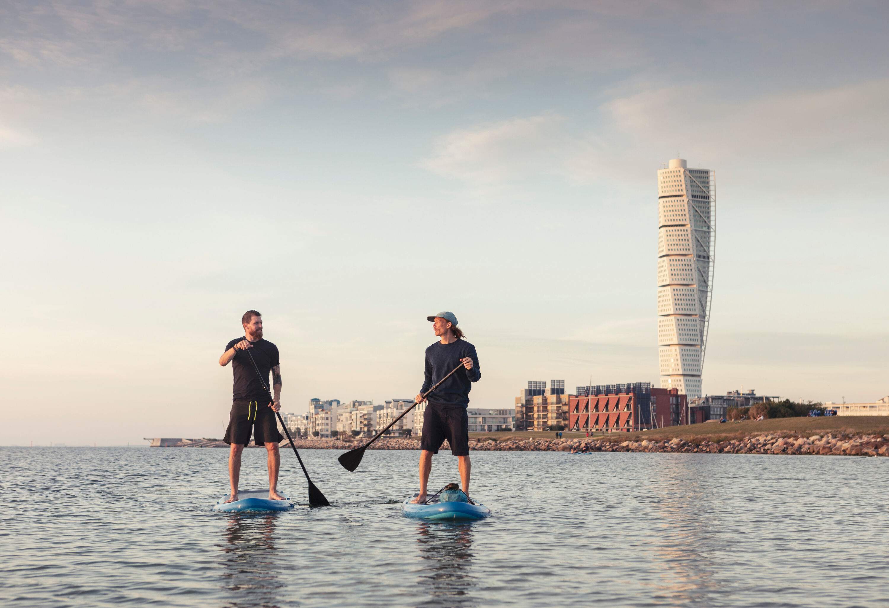 Two men stand on floating boards and paddle themselves to propel through the water while talking to each other with an iconic skyscraper in the background.