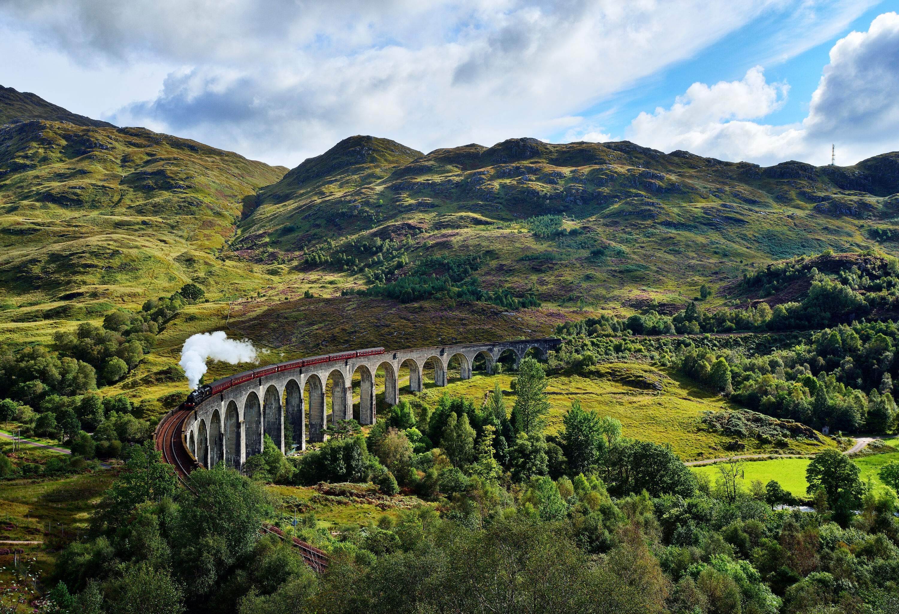 A steam train travels on a viaduct across a mountainous area covered in lush vegetation.