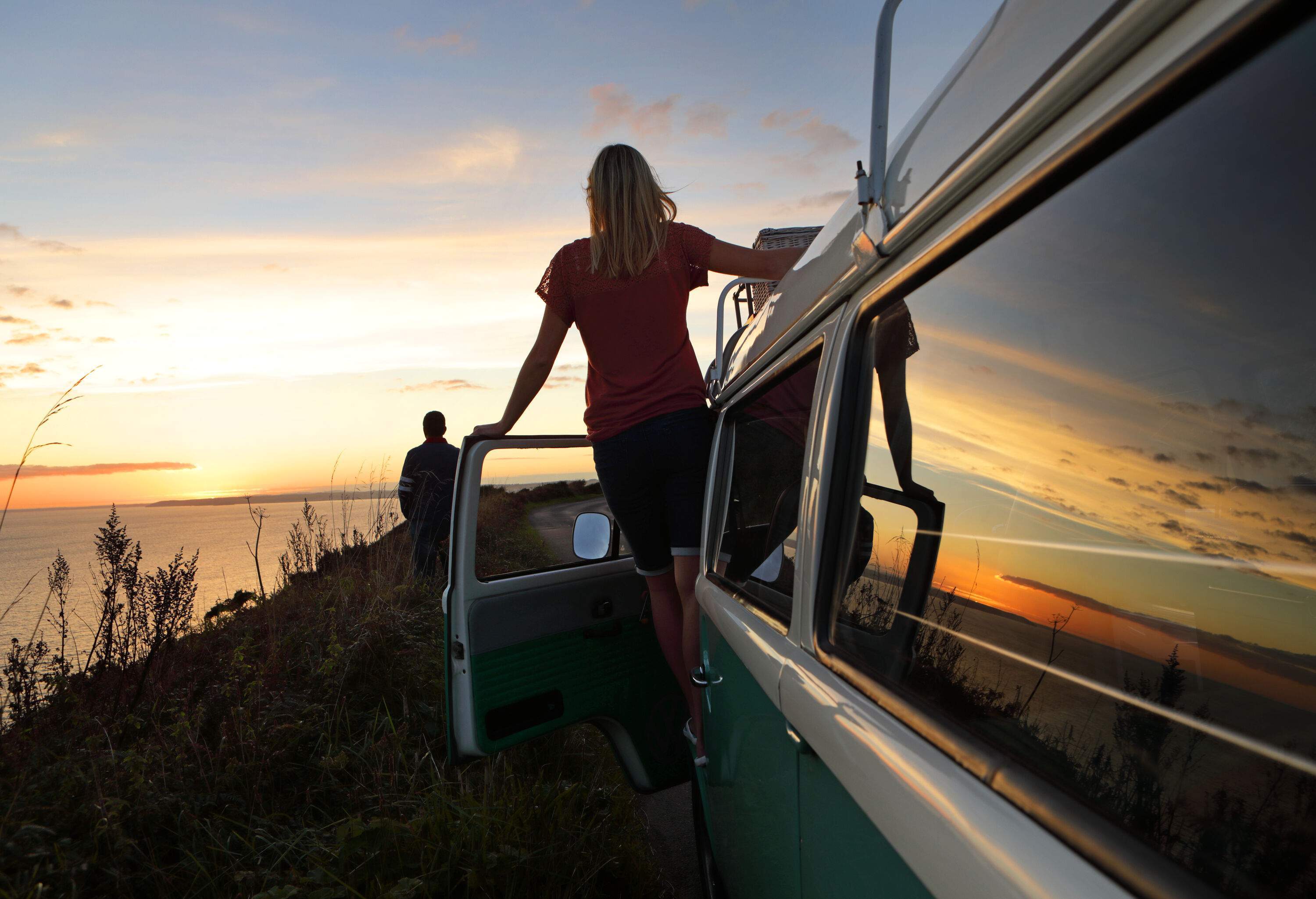 Silhouette of two people gazing at the sunset, one stands on the hillside and the other standing in the car with its door open.