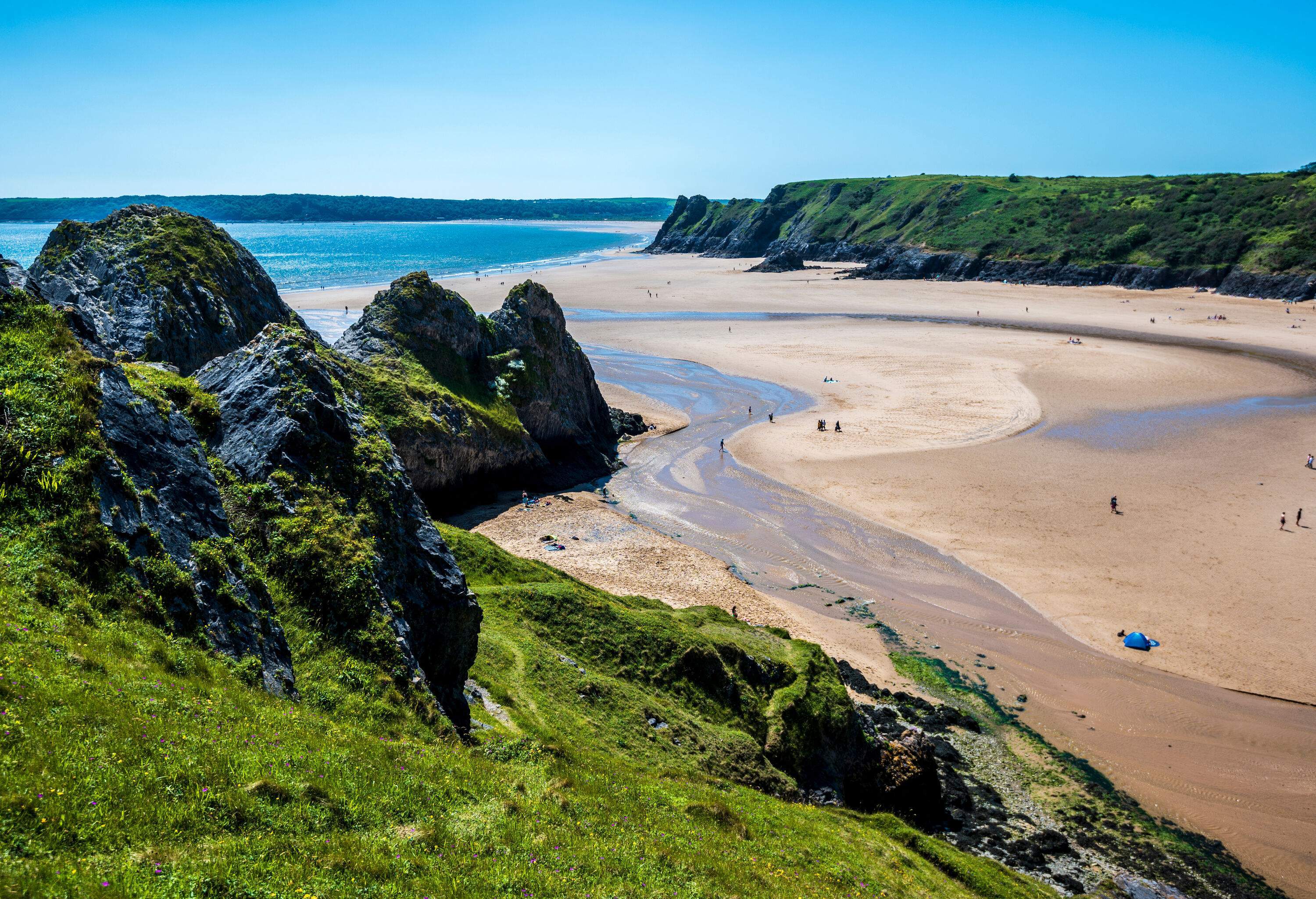 A vast sandy beach in the middle of rugged mossy cliffs with a stream flowing through the golden sand into the sea.