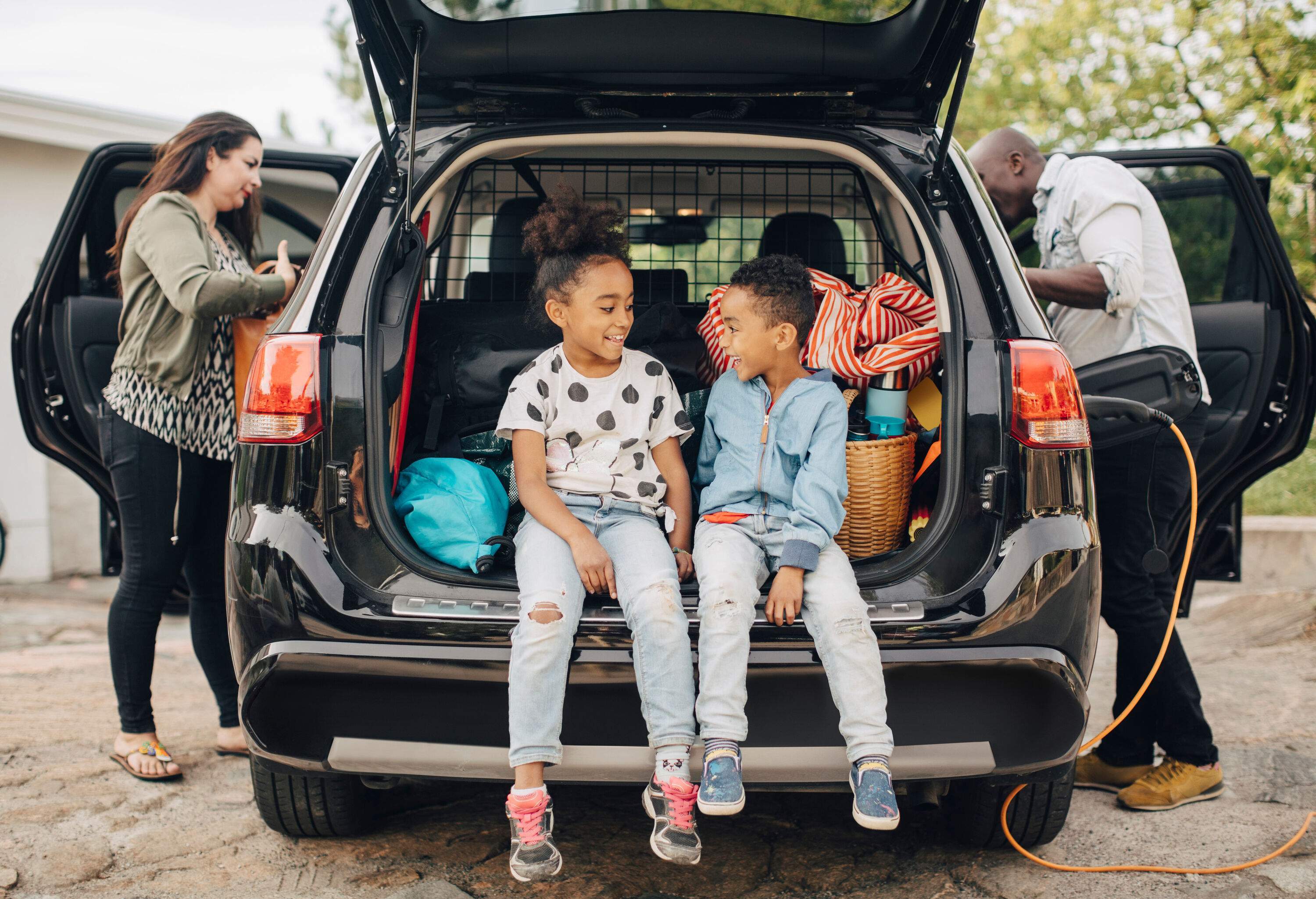 Happy siblings sit on a car trunk while their parents load up their bags inside the car.