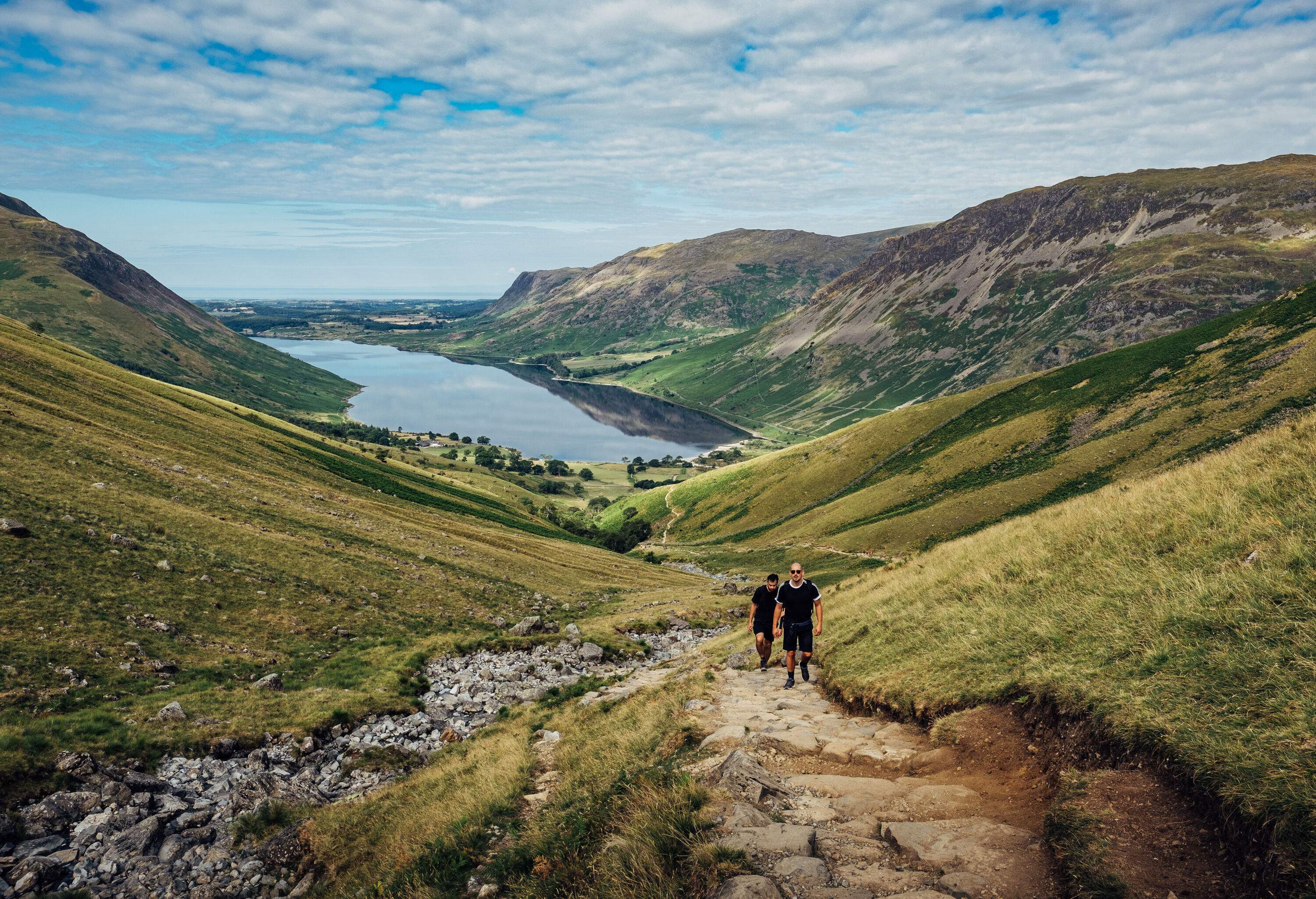 Two individuals ascending a stone staircase between two grass valleys with a lake in the distance.