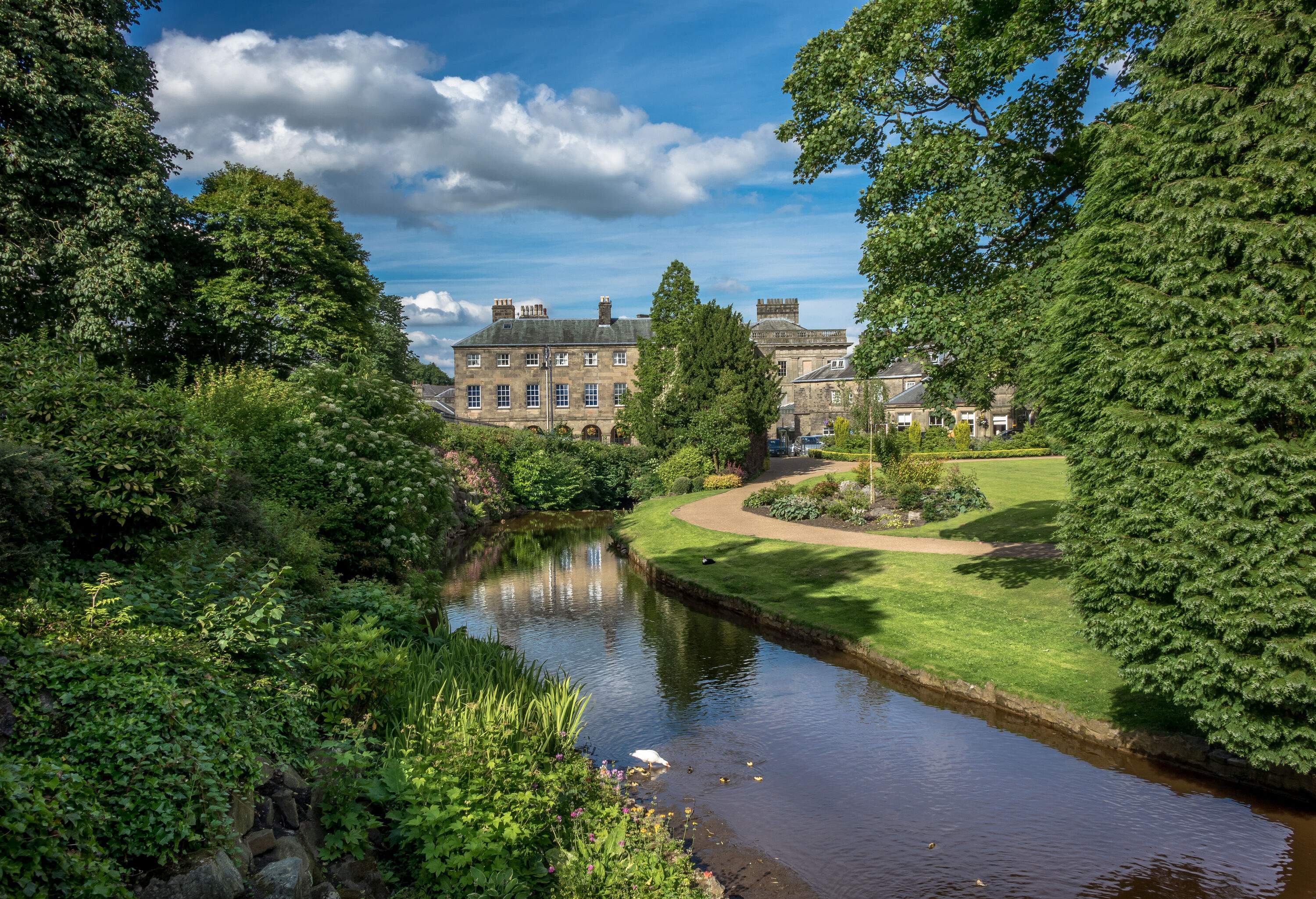 An old stone building is located at the end of a grassy path that winds through a pond surrounded by plants and trees.