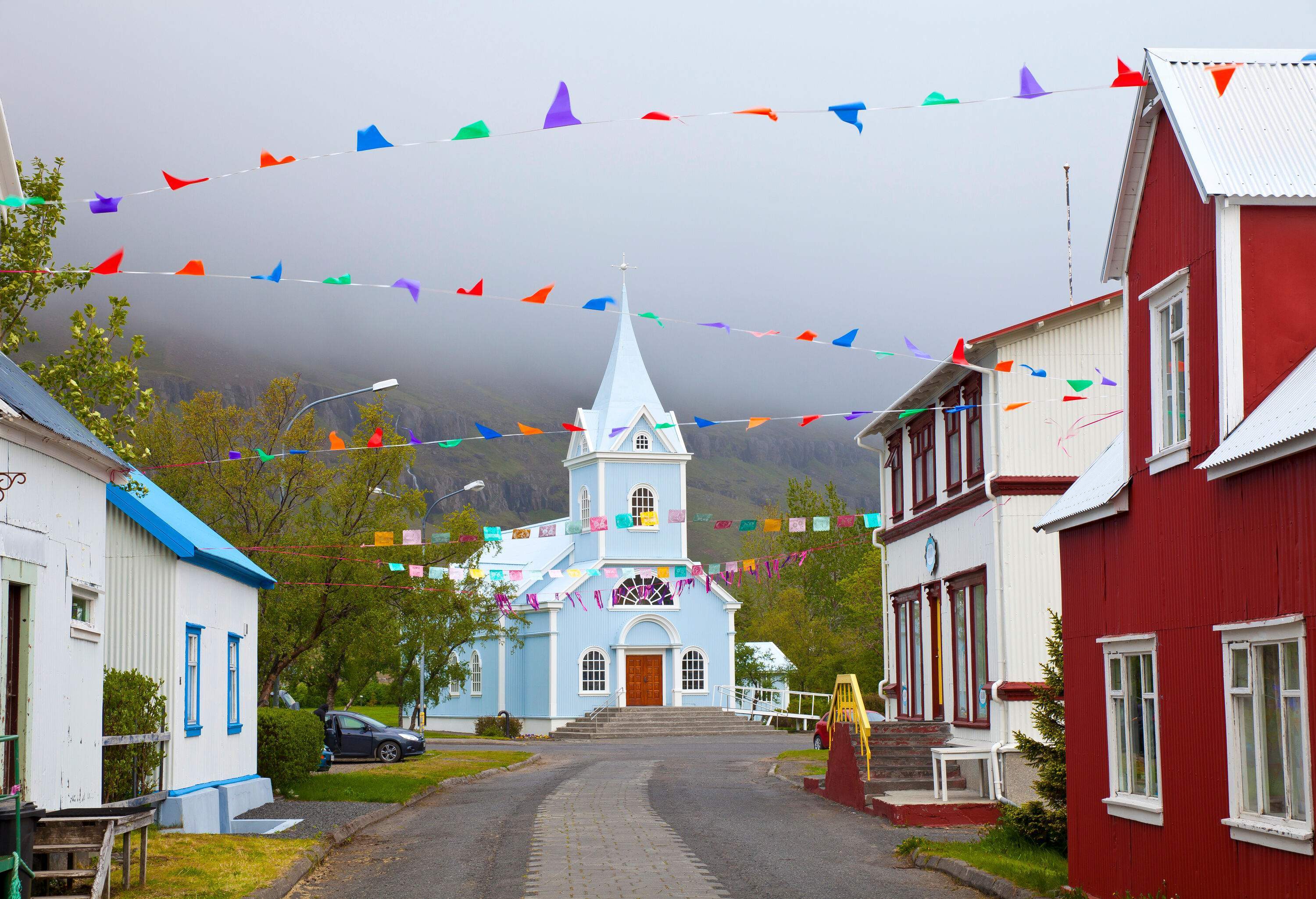 A sky-blue church on the mountainside at the end of an asphalt road in the middle of colourful houses on a foggy day.