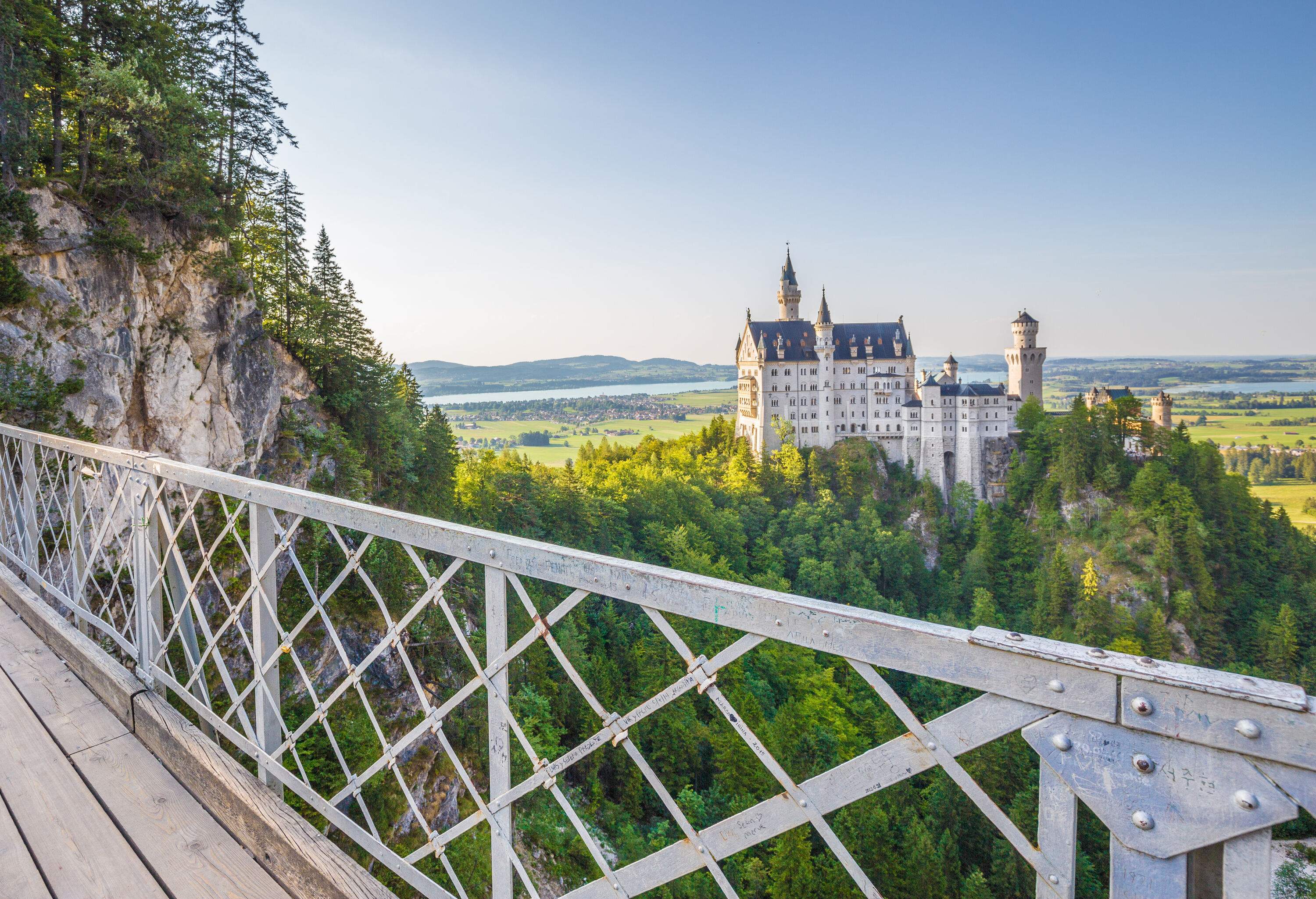 The Romanesque Revival palace of the famous Neuschwanstein Castle is surrounded by lush forest seen from the bridge.