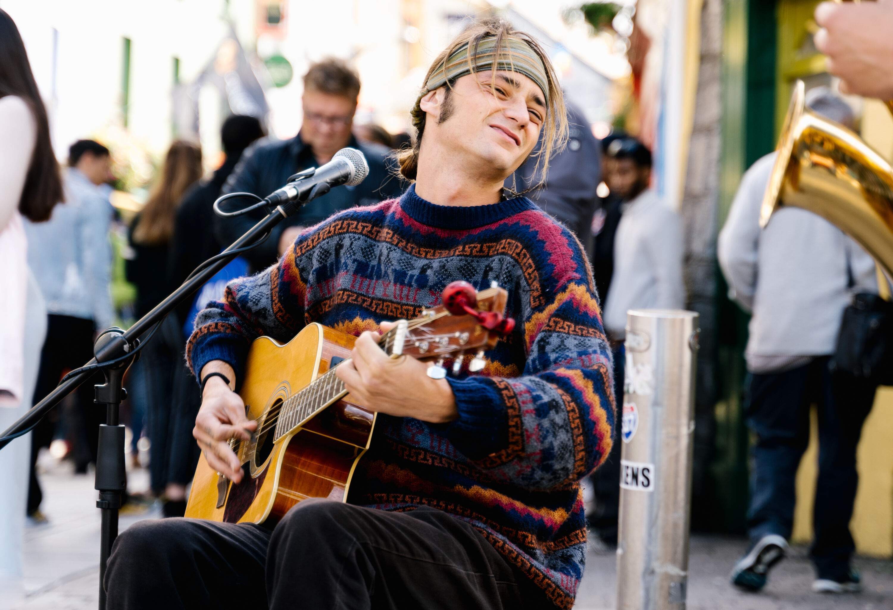 A talented musician sits on a street, playing a guitar in front of a microphone, wearing a colourful cardigan and bandana, captivating passersby with soulful melodies.